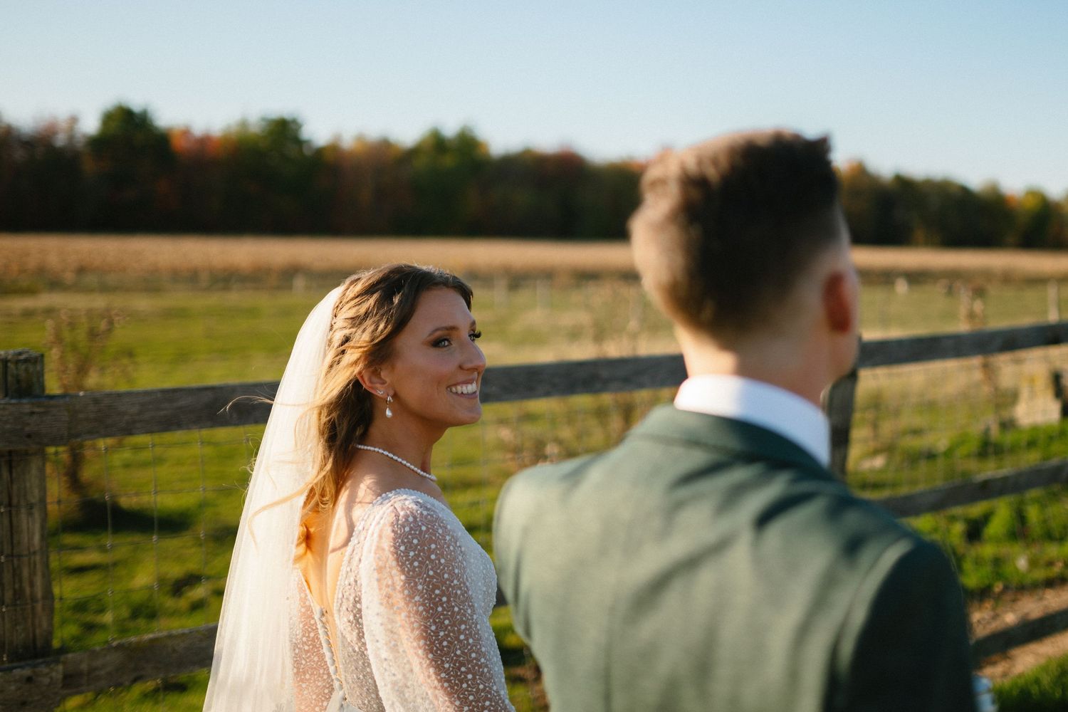Newlyweds stand together against rustic wooden fence at sunset.