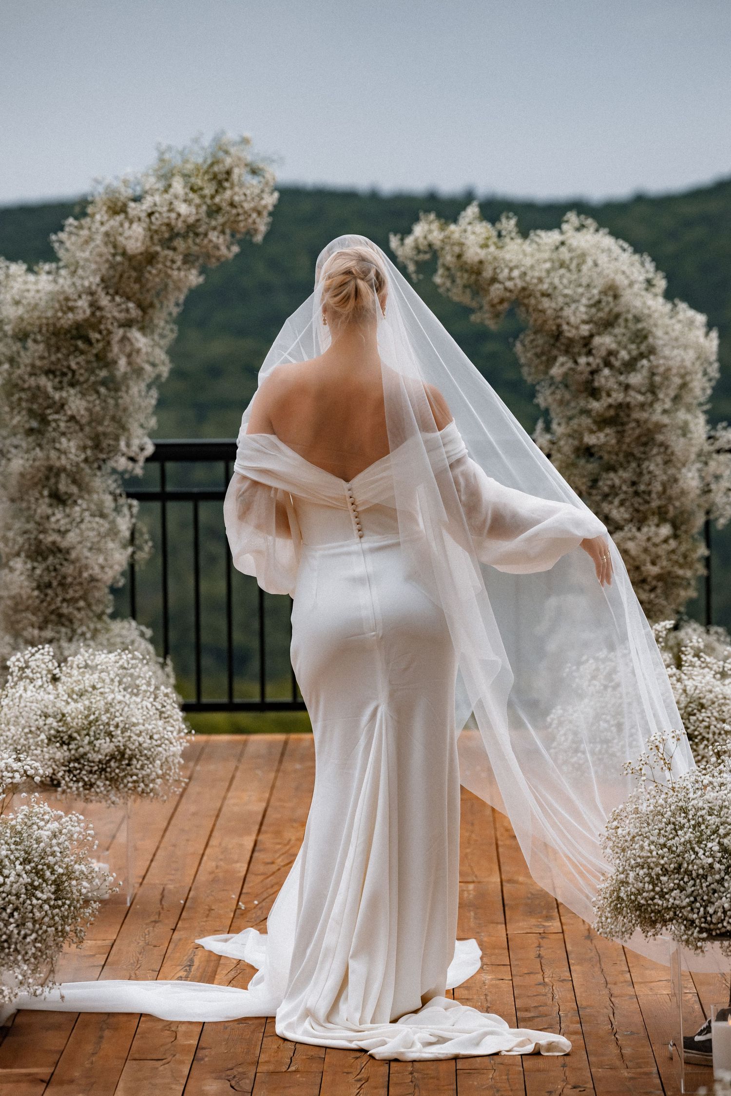 Elegant white wedding dress with flowing veil against mountain backdrop on wooden deck.