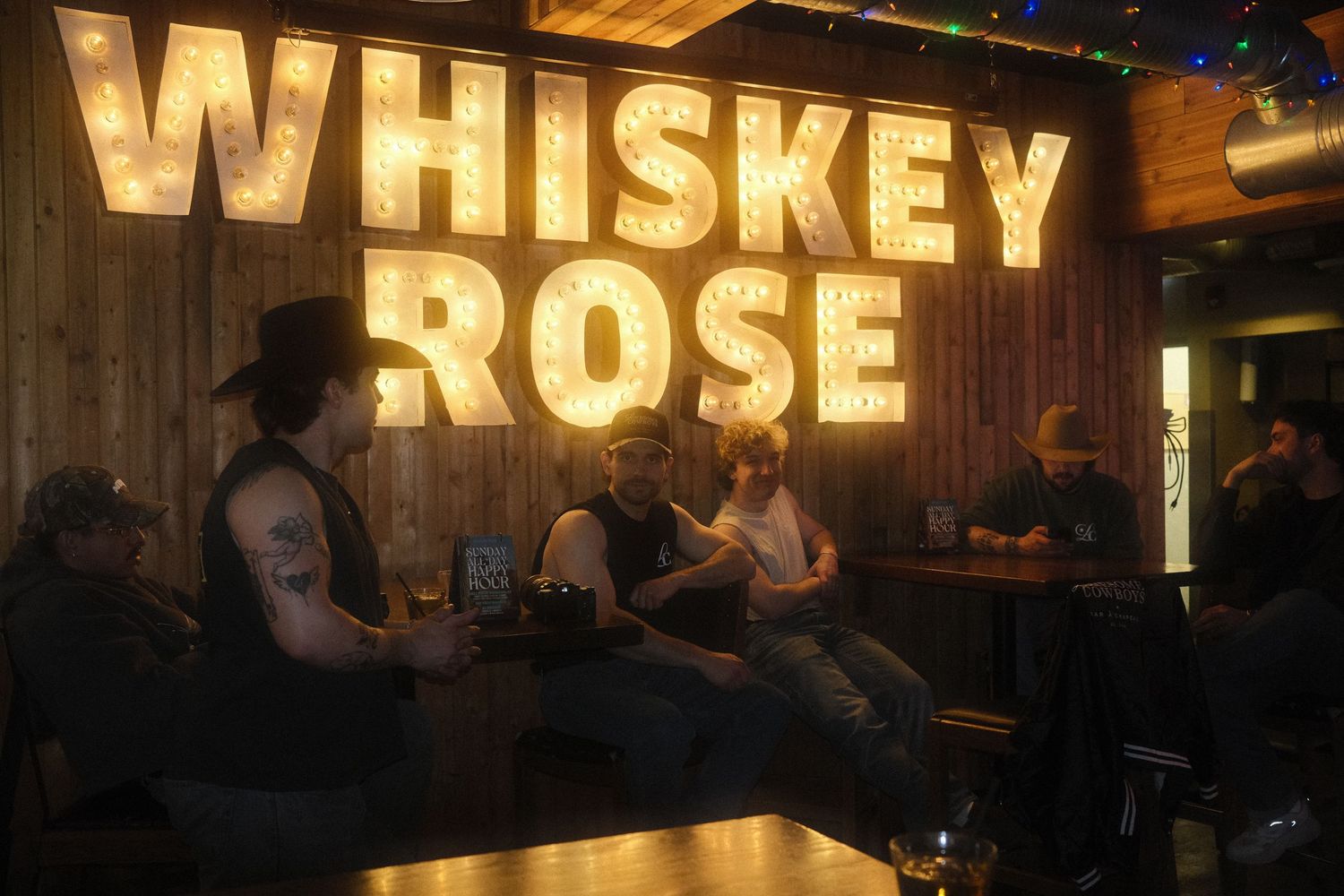 People socializing inside Whiskey Rose bar with rustic wood walls and illuminated marquee sign lighting.