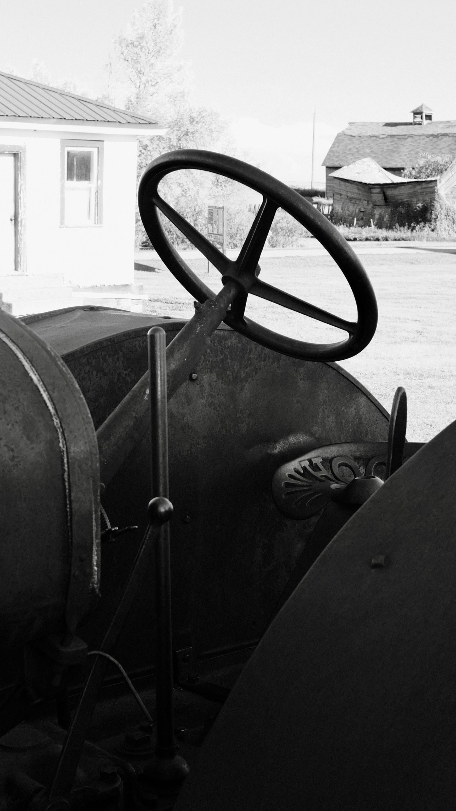 Black and white photo of a vintage tractor steering wheel with farm buildings in the background.