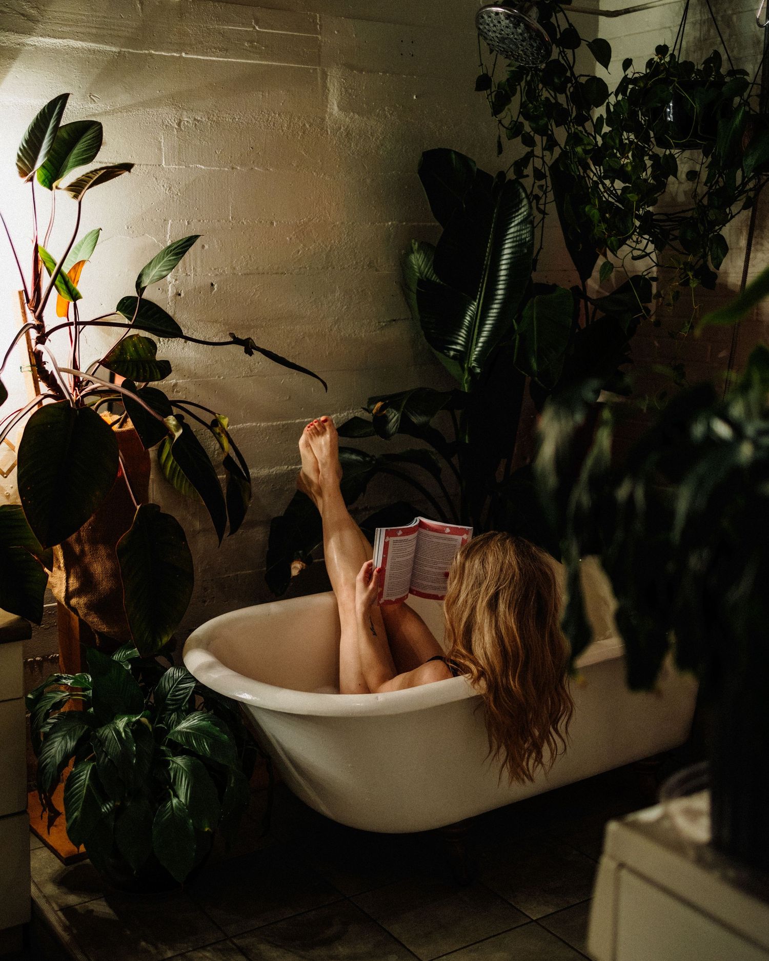 Person relaxing in bathtub surrounded by indoor plants reading a book.