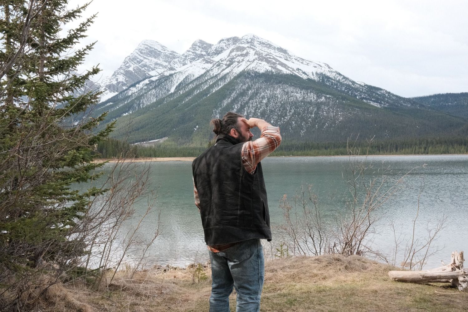 Person standing by a mountain lake with snow-capped peaks and evergreen trees in the background.