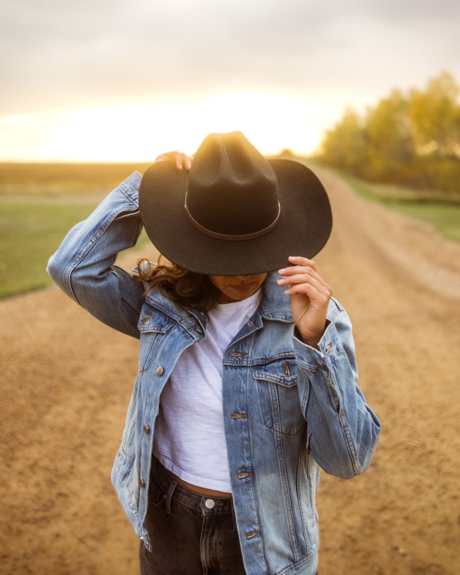 Woman in denim jacket and black cowboy hat standing on a rural dirt road at golden sunset.