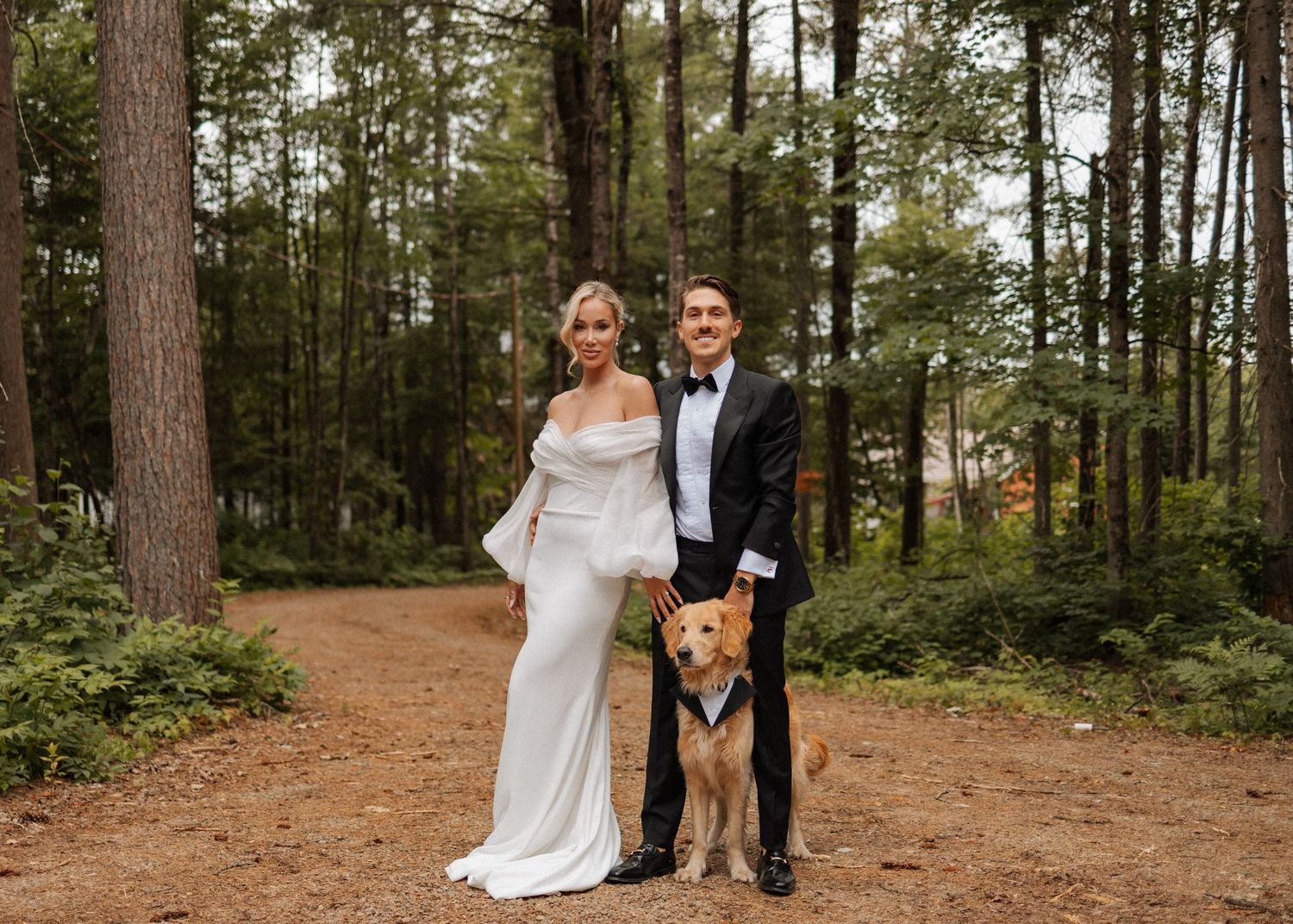 Couple in wedding attire posing on a forest path surrounded by tall pine trees.