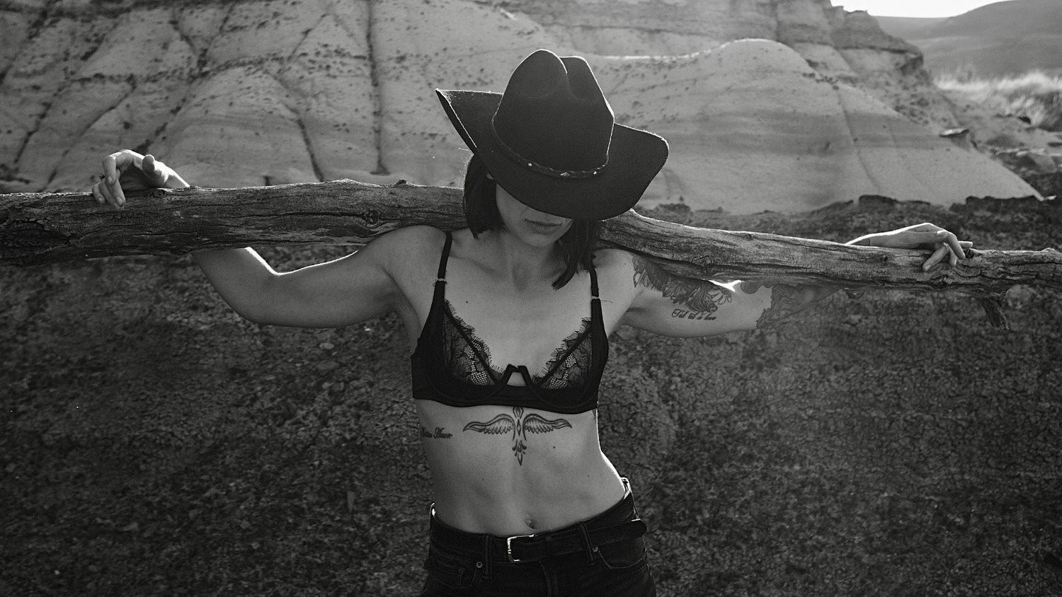 Black and white photo of a woman in a cowboy hat and lace bralette posing with a wooden beam in a desert landscape.