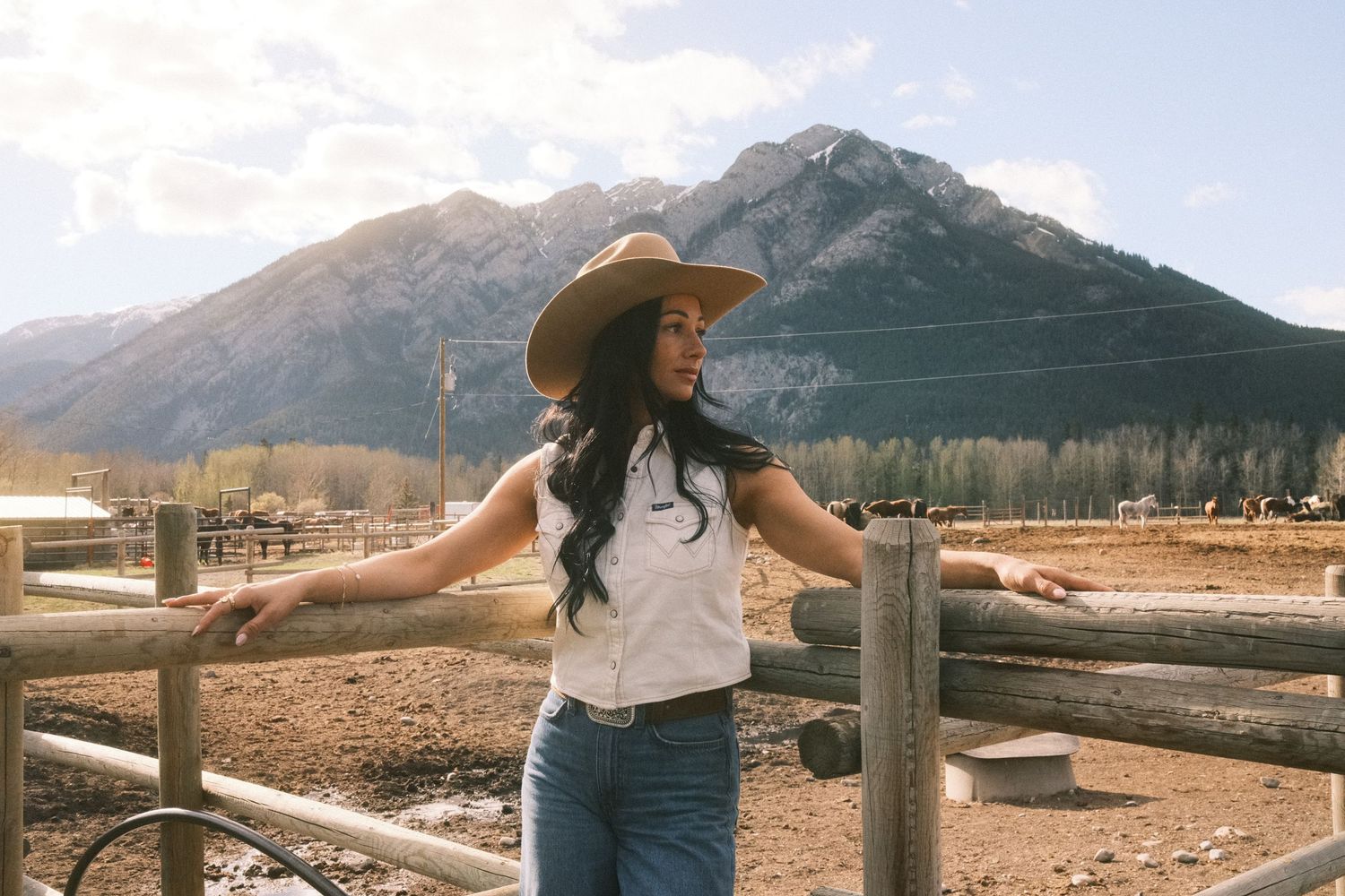 A young woman in a cowboy hat and jeans poses by a wooden fence with a mountain backdrop.