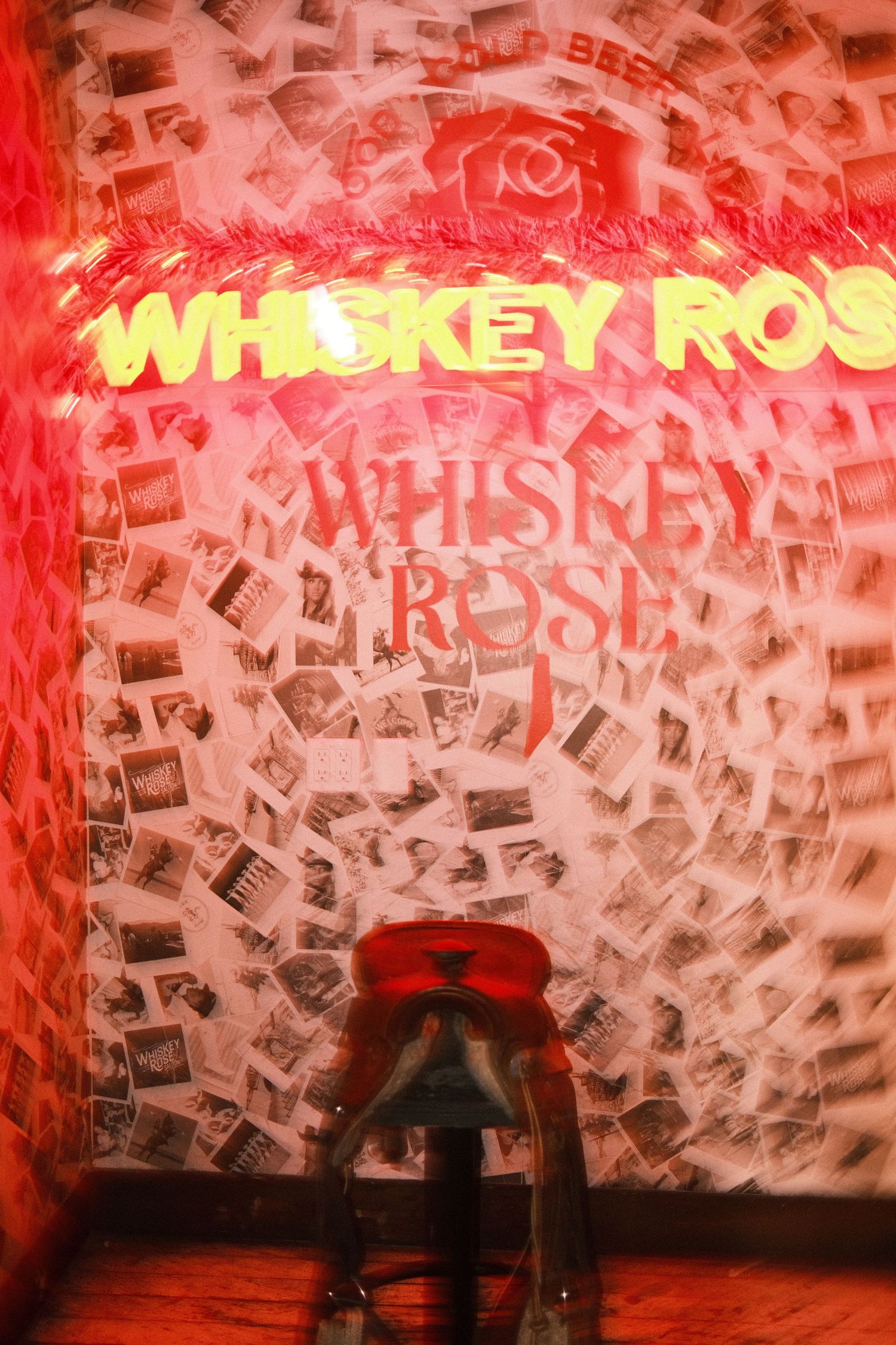 Whiskey Rose bar neon sign glowing red above a barstool, surrounded by polaroid photos on the wall.