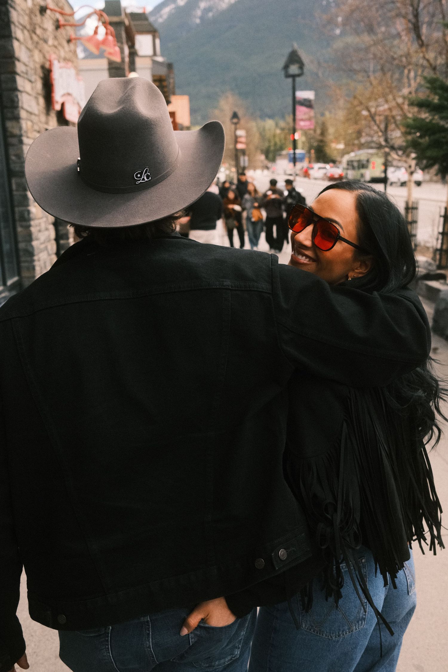 Couple walking down a mountain town street, woman in red sunglasses and fringe jacket smiling at man in black cowboy hat.