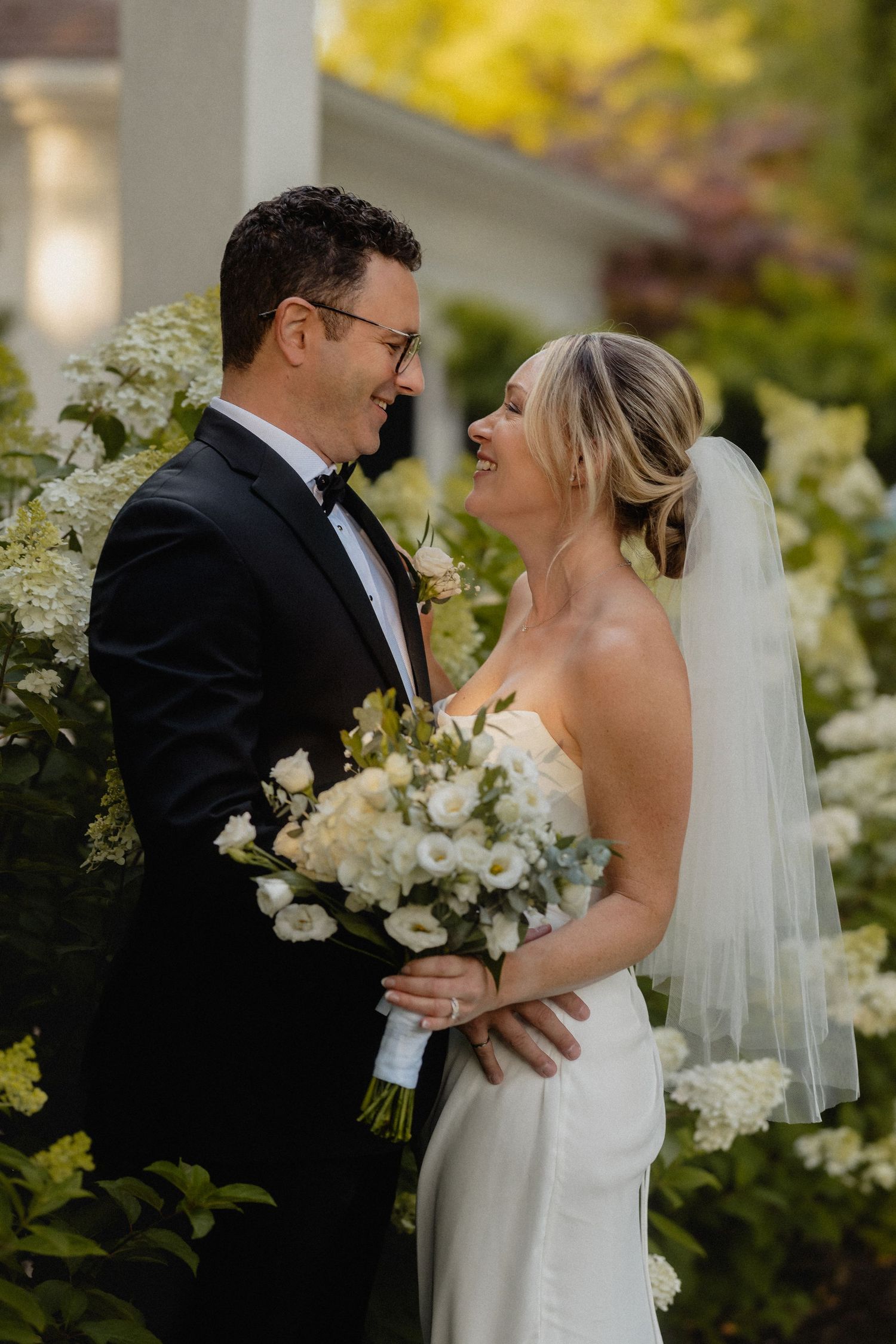 Joyful wedding portrait with couple laughing among white hydrangeas and greenery.