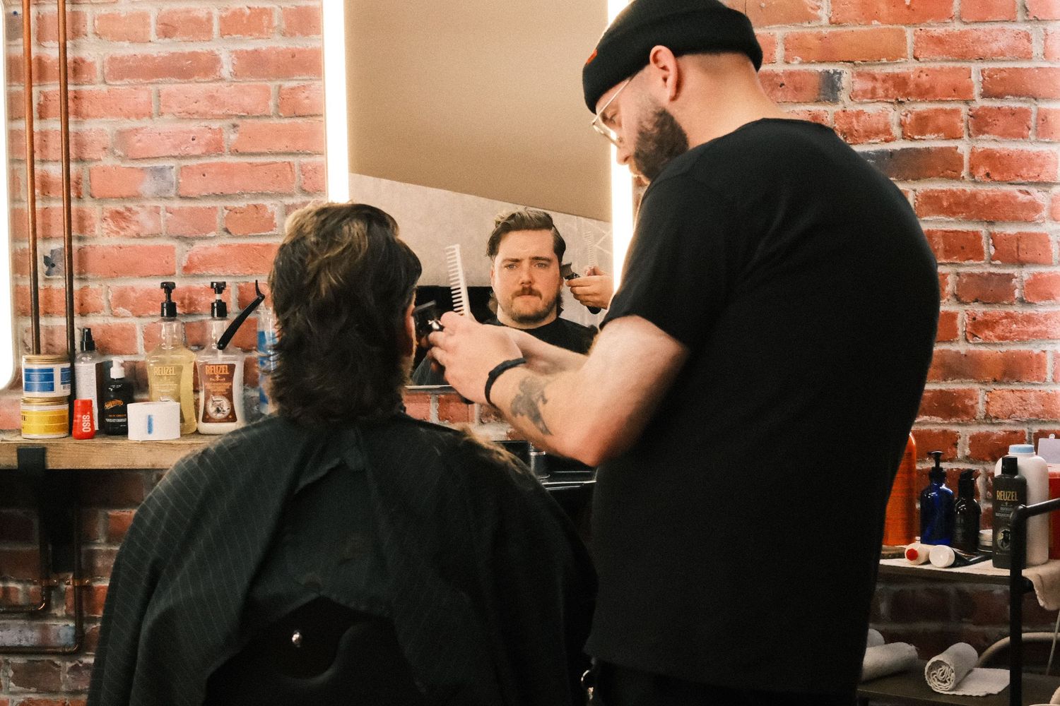 A barber in a black cap cuts a client's hair in a rustic brick-walled barbershop with mirrors and grooming products.