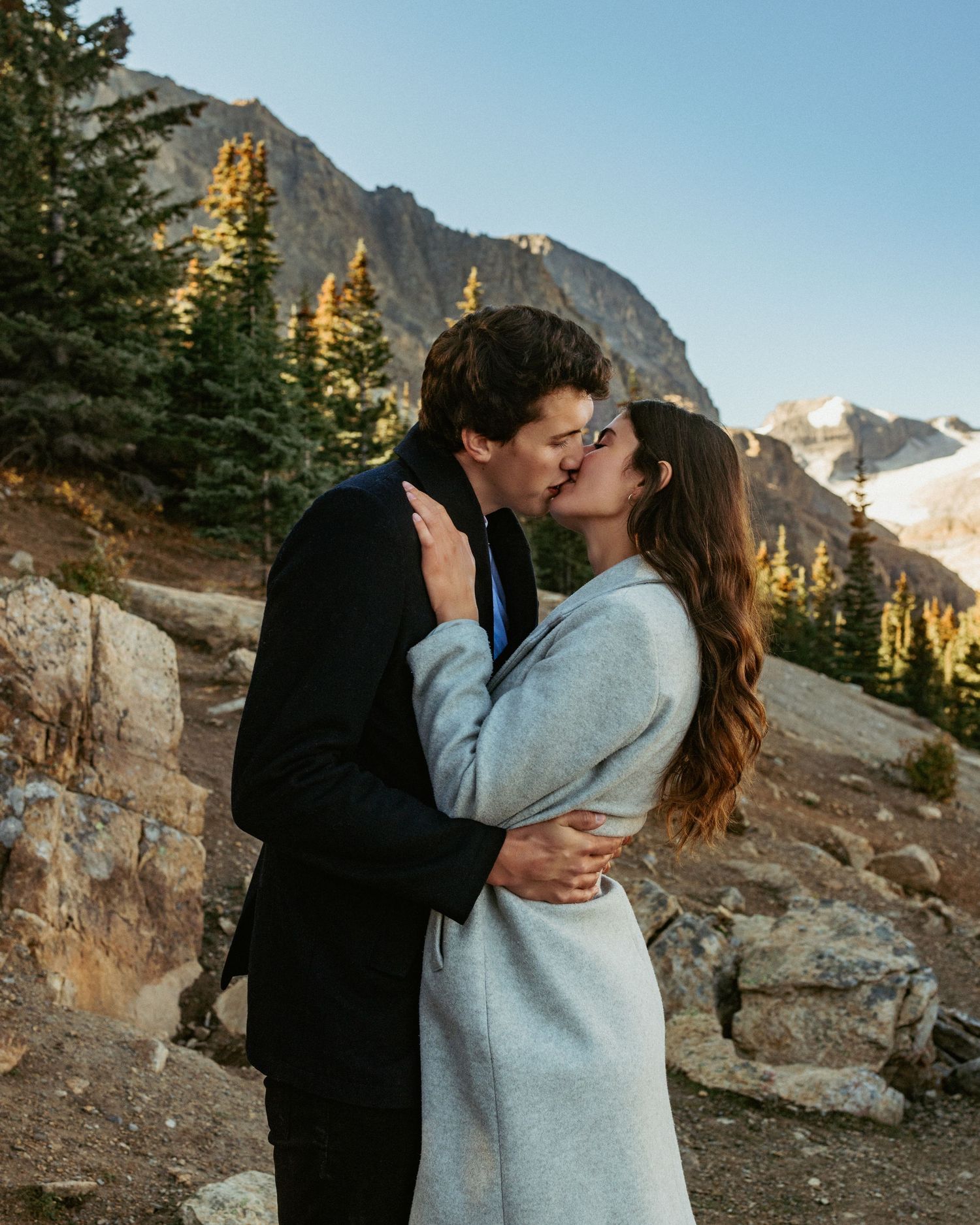 Couple embracing on mountain overlook with dramatic rocky peaks and golden sunlight in background.