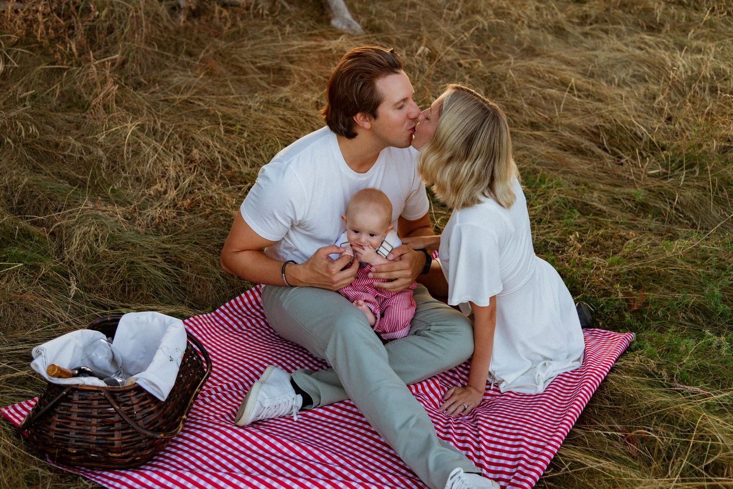 Family having a picnic on a red checkered blanket outdoors with their baby.
