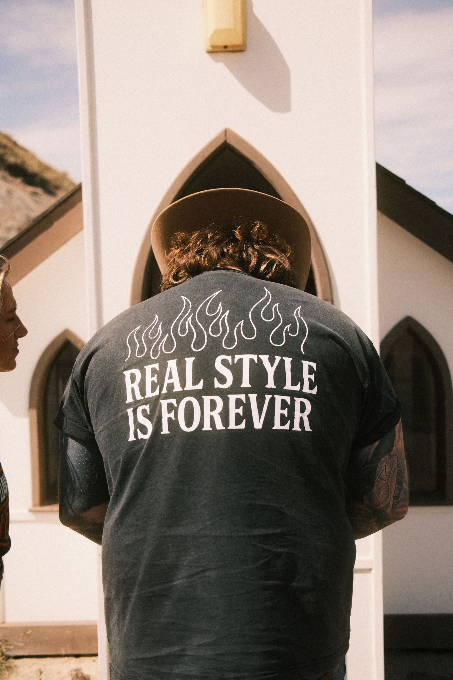 Person wearing dark graphic tee with flames and 'Real Style Is Forever' text, standing outside a white church.