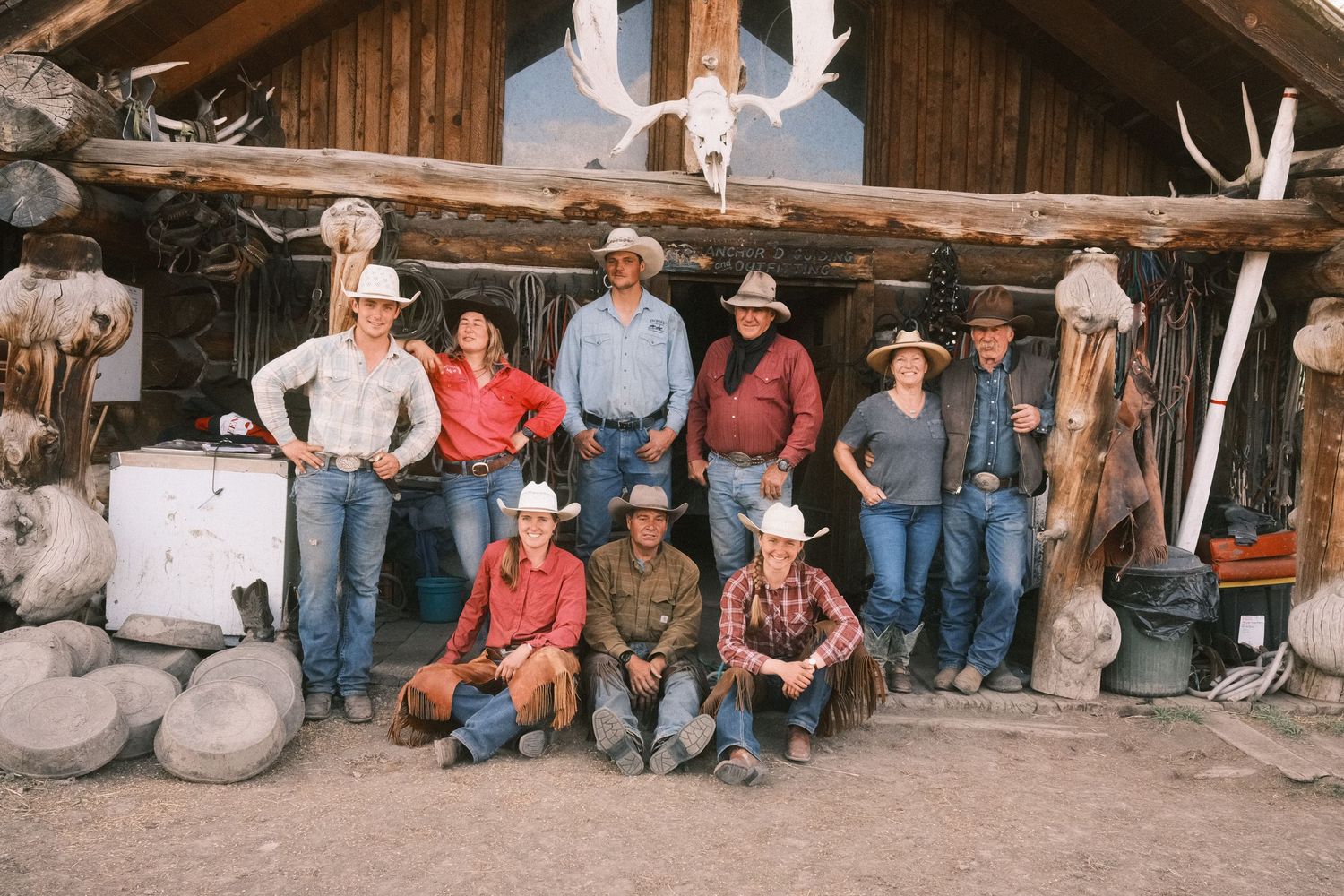 Group of cowboys and cowgirls posing in front of a rustic log cabin with antlers and western decor.