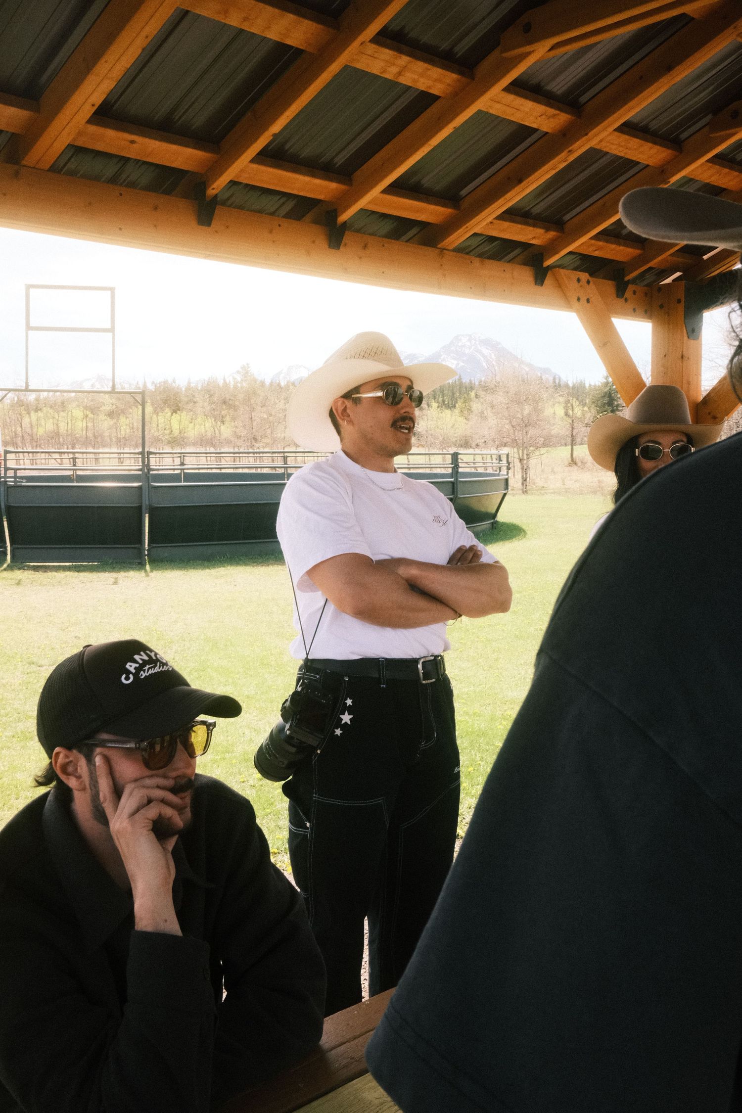 Cowboys in hats gather under a wooden pavilion at a ranch, with green fields and mountains in the background.