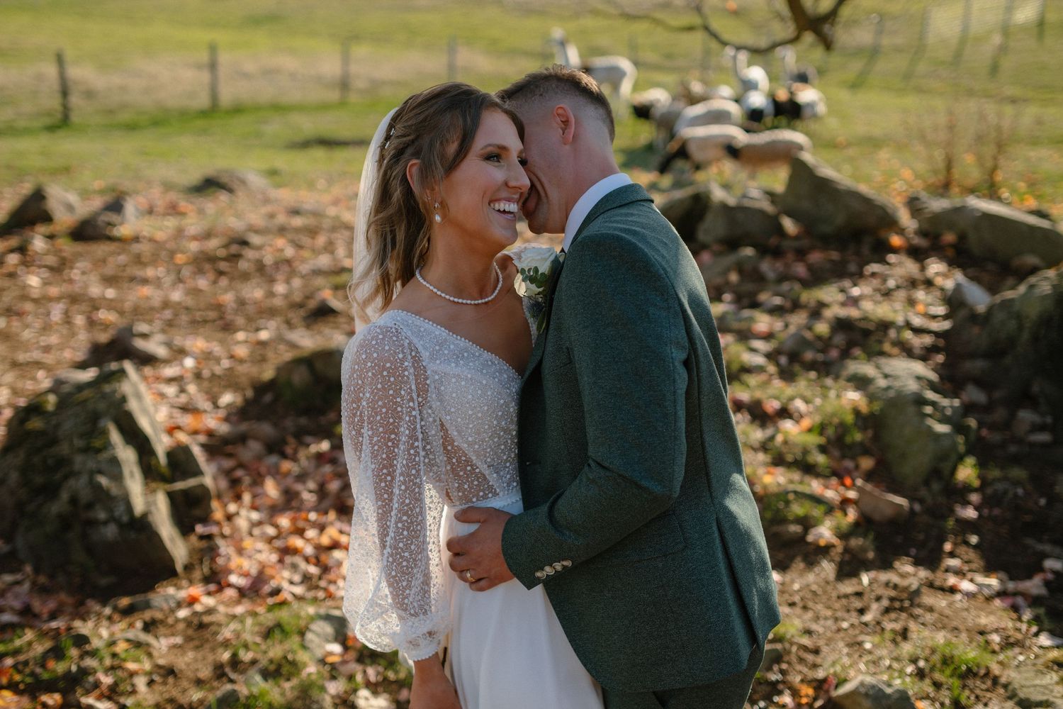 Wedding couple embraces in sunlit field with stone wall backdrop.