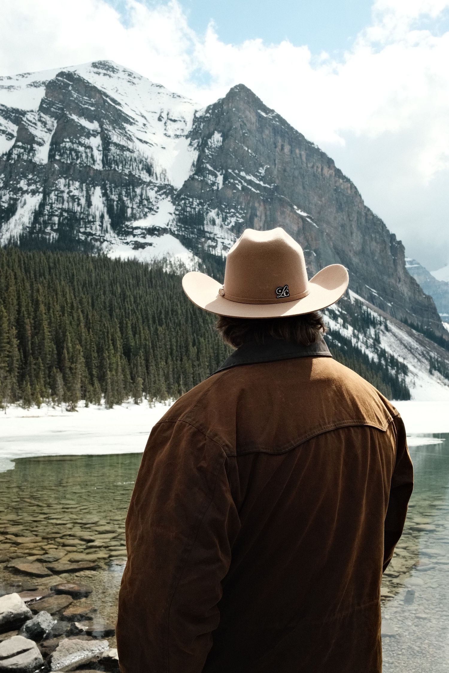 A man in a cowboy hat and brown jacket gazes at a stunning mountain lake with snow-capped peaks and pine forests.