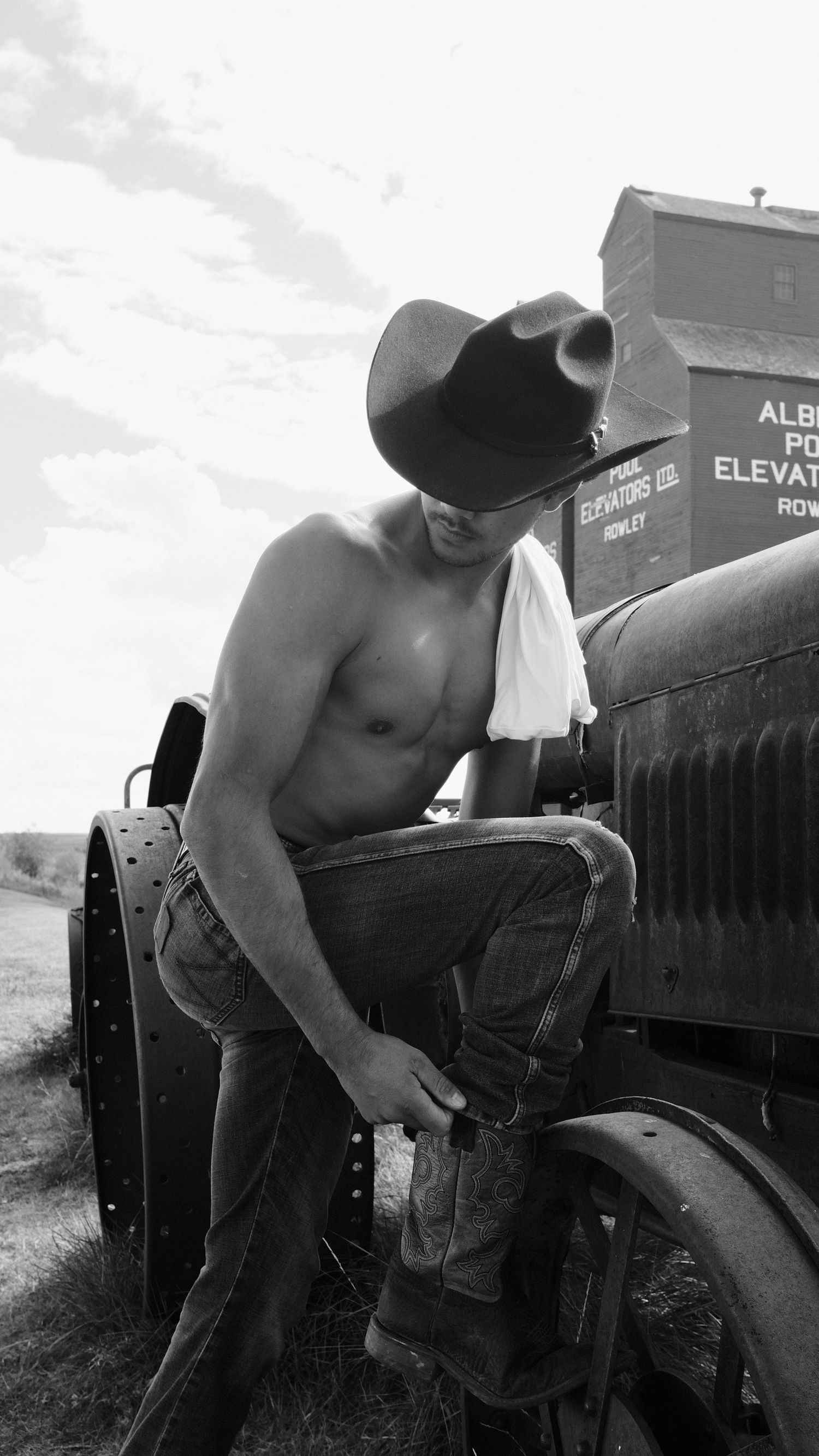 Shirtless cowboy in hat leans against vintage black truck in rural setting, black and white photography.