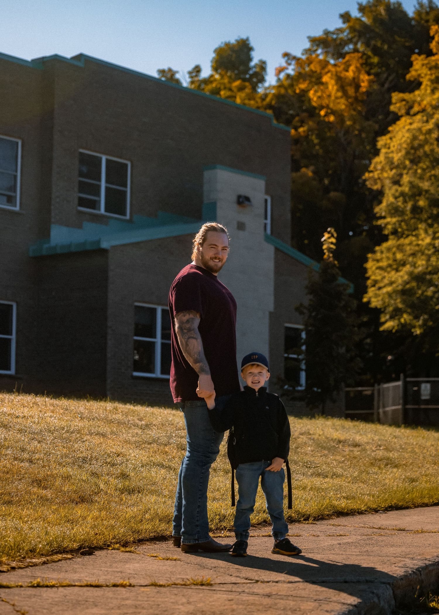 A tattooed man and young boy hold hands outside a brick building on a sunny autumn day.