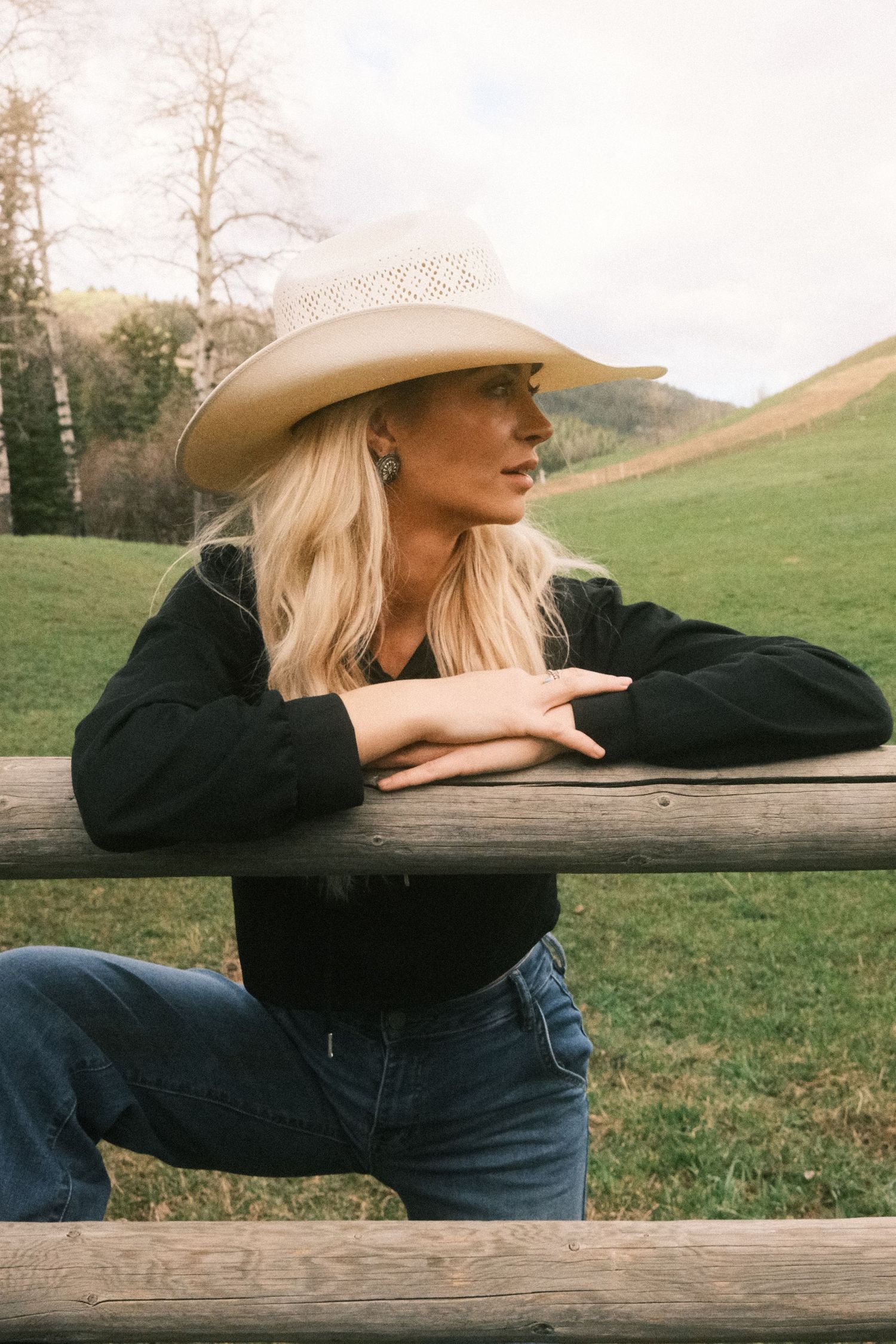 Blonde woman in cowboy hat and black shirt leaning on wooden fence in green countryside setting.