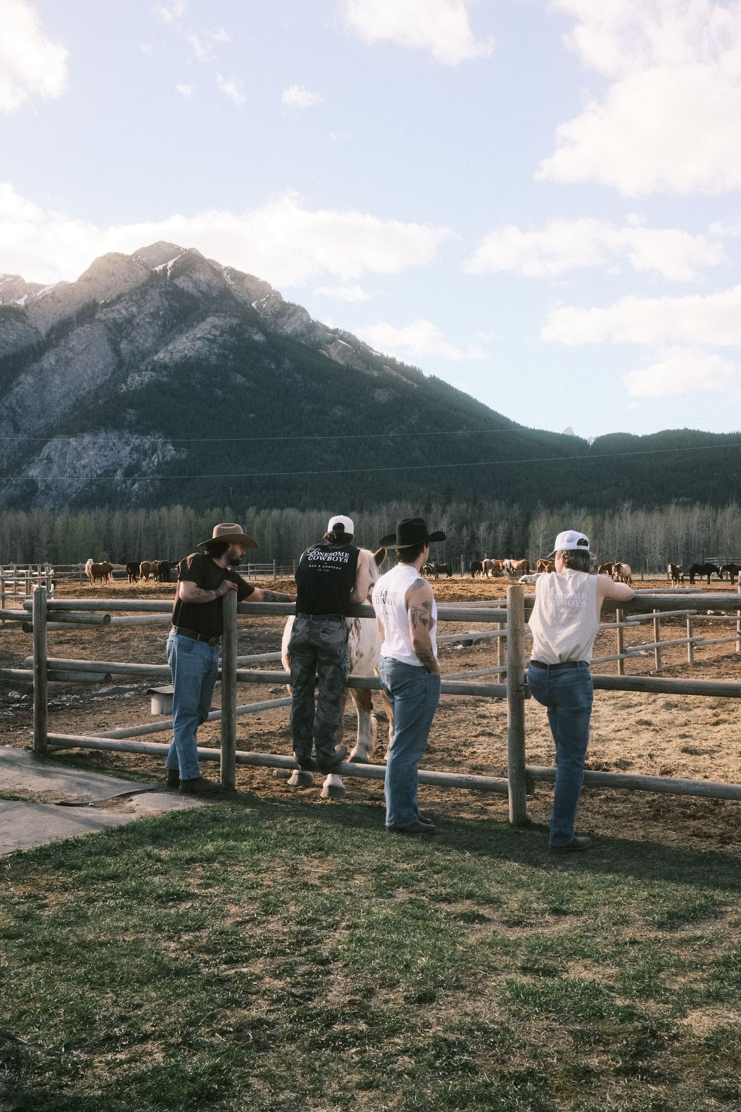 Four cowboys leaning on a wooden fence overlooking a ranch with horses and mountains in the background.