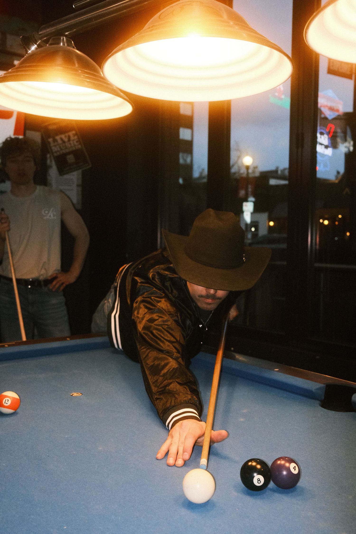 Man in cowboy hat playing pool, leaning over blue billiard table with cue stick under bright hanging lights.