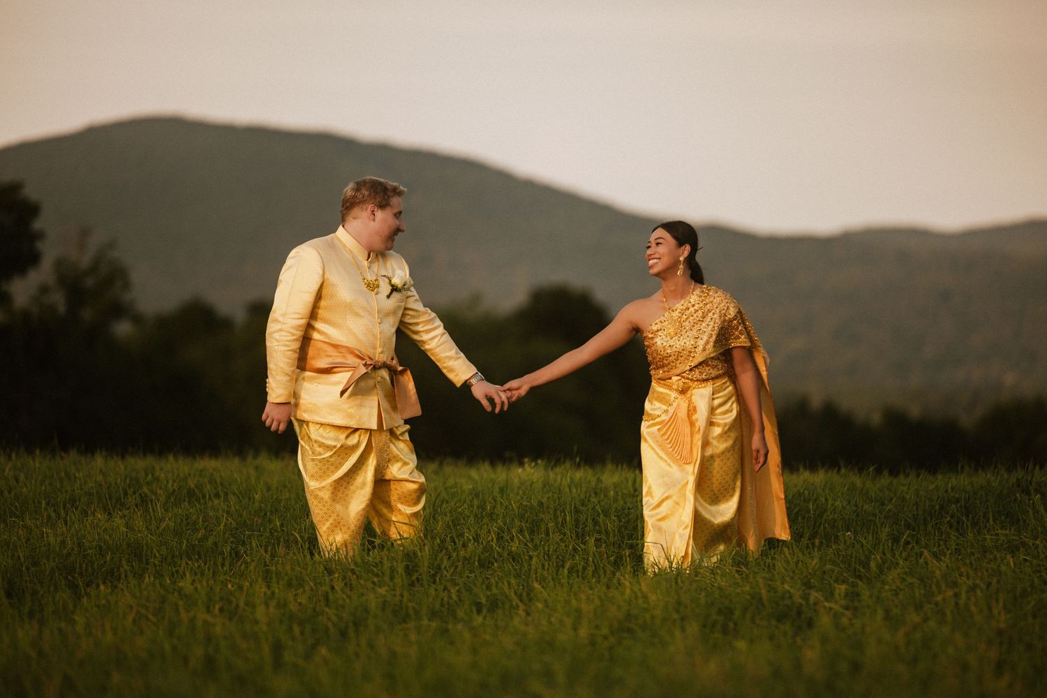 Couple in cream-colored outfits walking through grassy field at sunset.