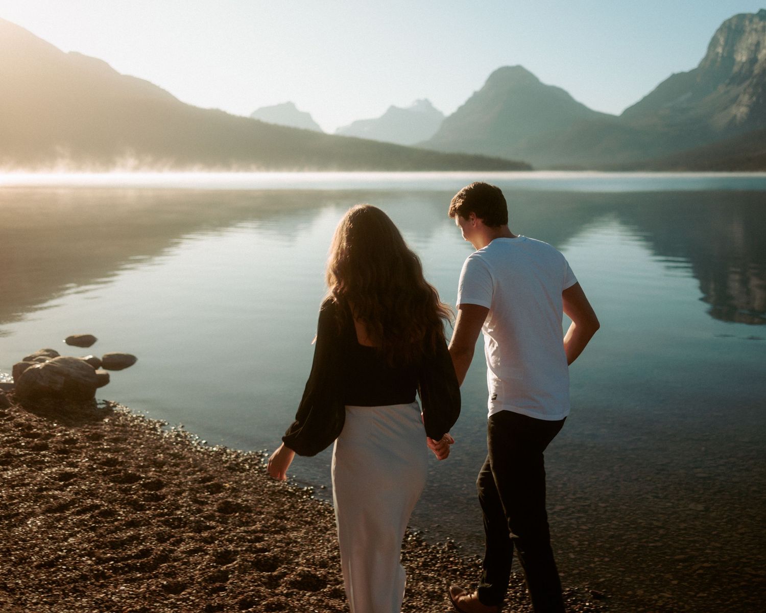 Couple exploring a rocky lakeshore with majestic mountains in the background.
