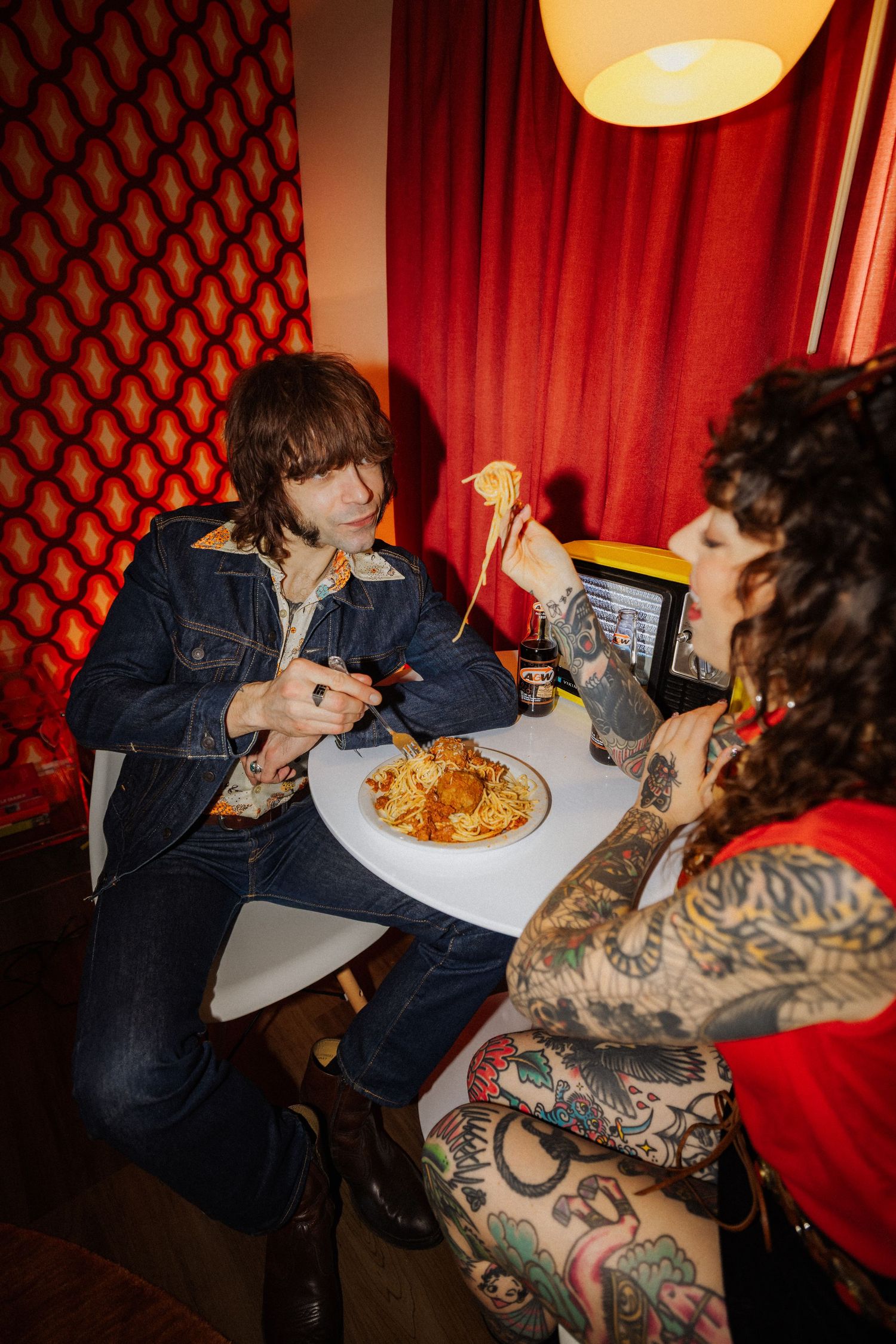Two people with tattoos sharing pasta at a restaurant with red decor.