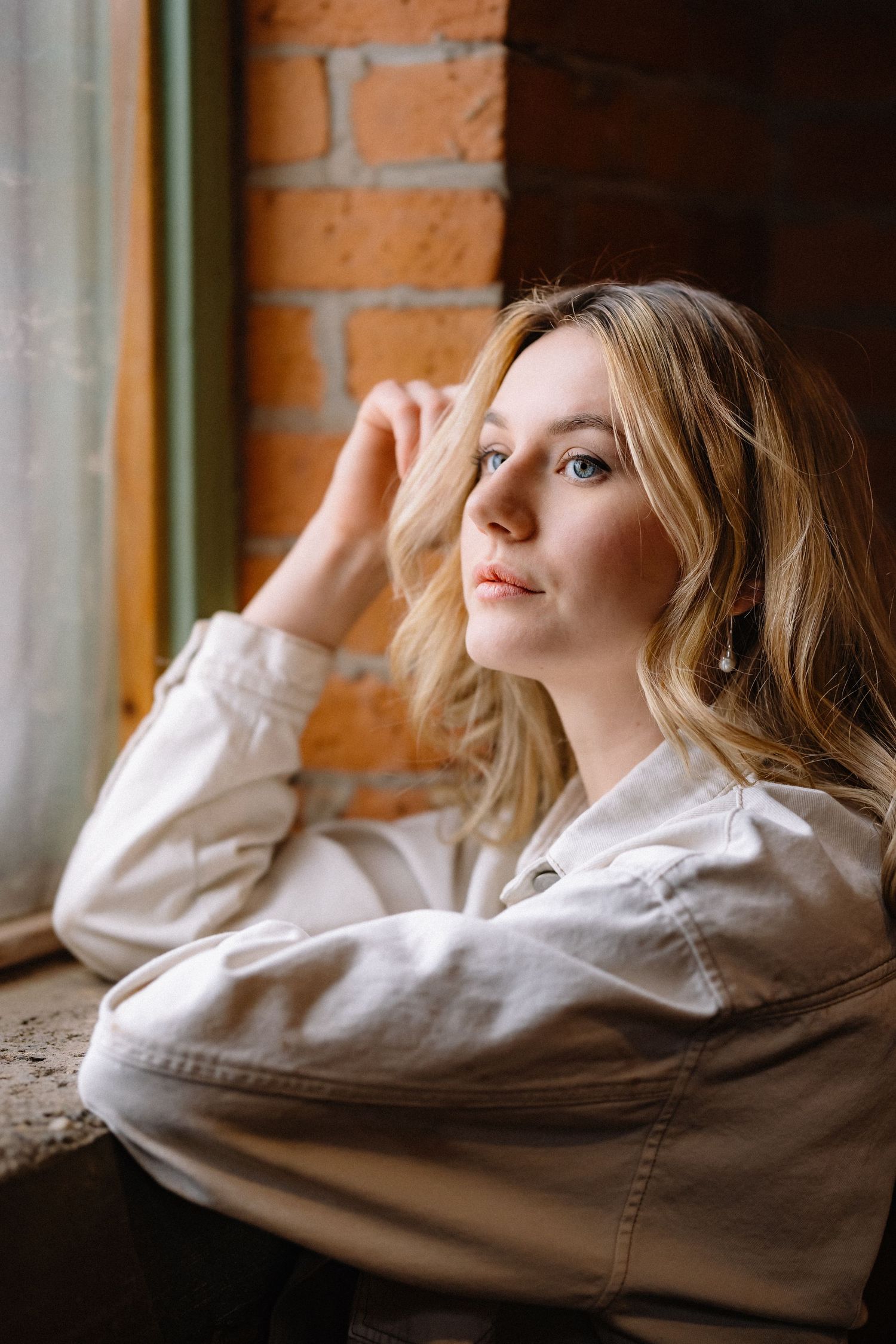 Person in beige hoodie relaxing by window with exposed brick wall.