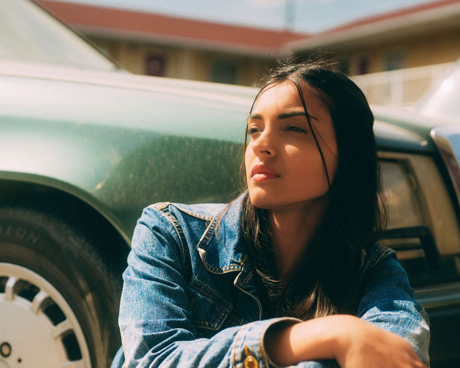 Person in denim jacket lounges against vintage car in natural lighting.