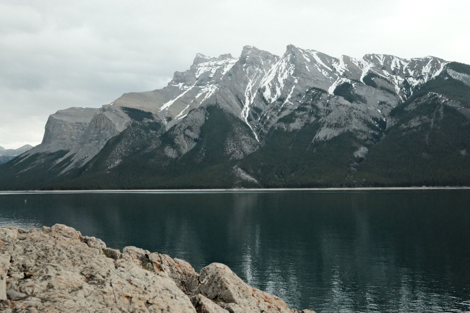 Snow-capped mountain range reflected in calm blue lake waters with rocky shoreline in Banff National Park, Canada.