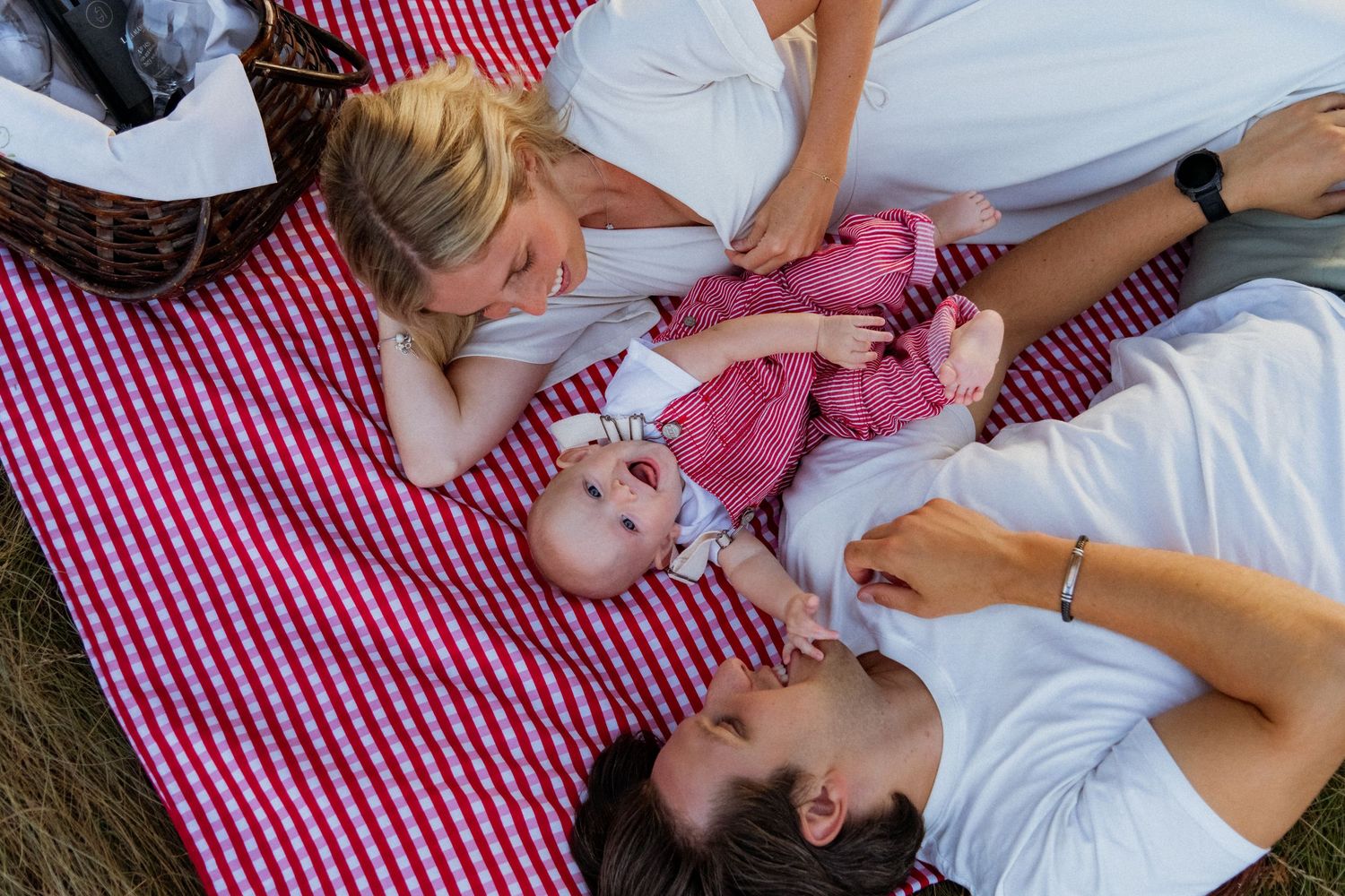 Family lying on a red checkered picnic blanket playing with their baby.
