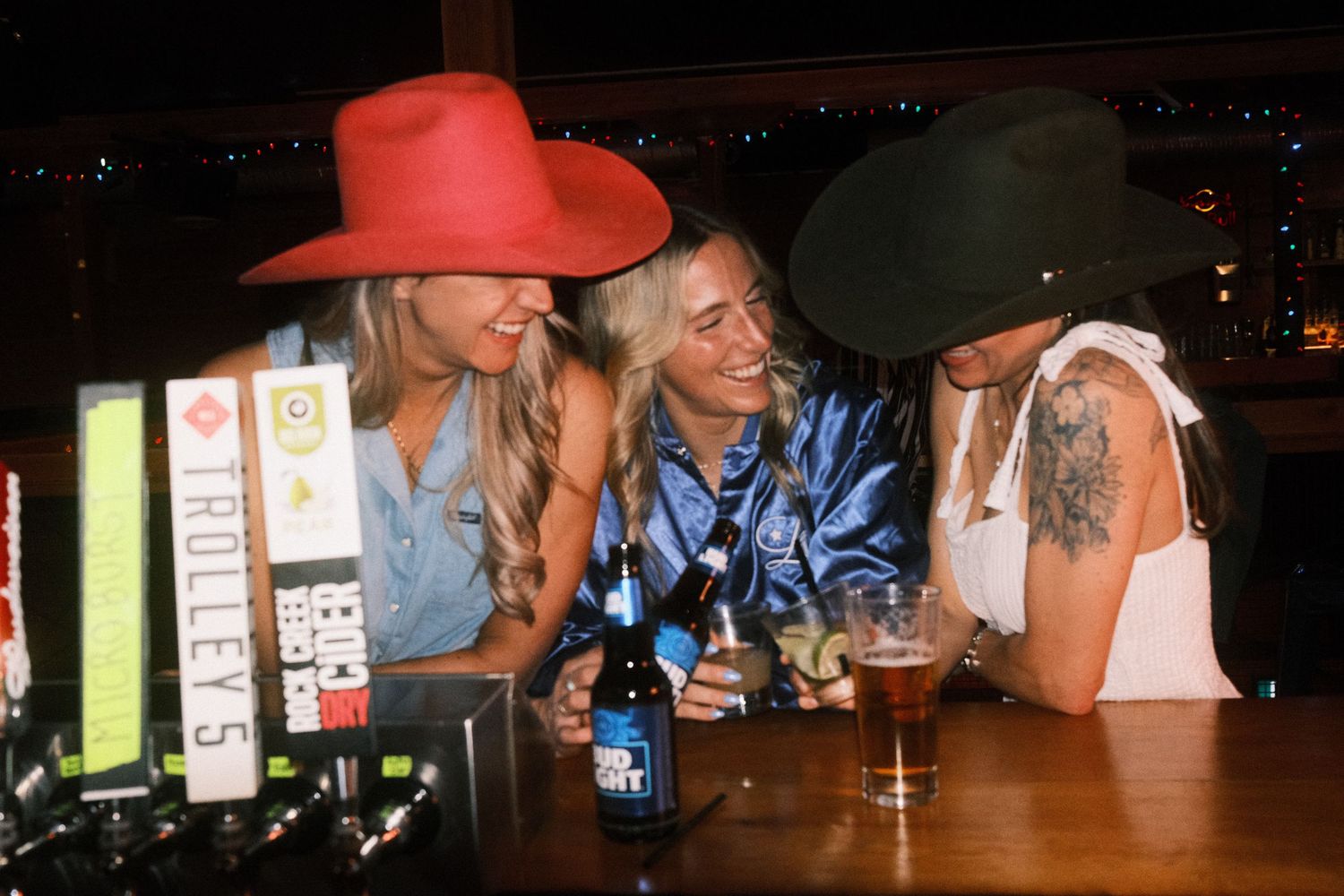 Three women in cowboy hats laughing together at a bar with drinks on the counter.