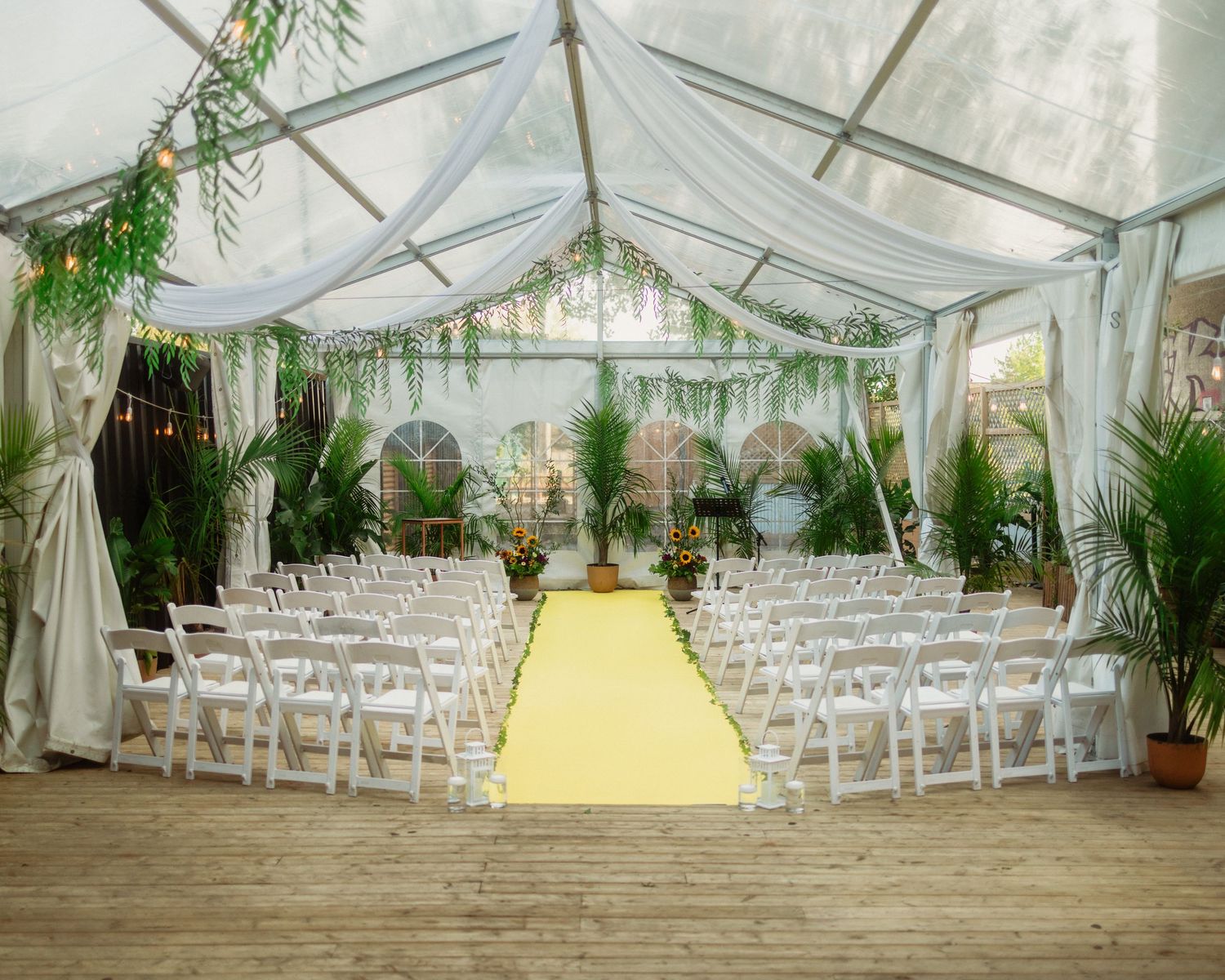 White wedding ceremony setup under a tent with rows of chairs and tropical plant decorations.