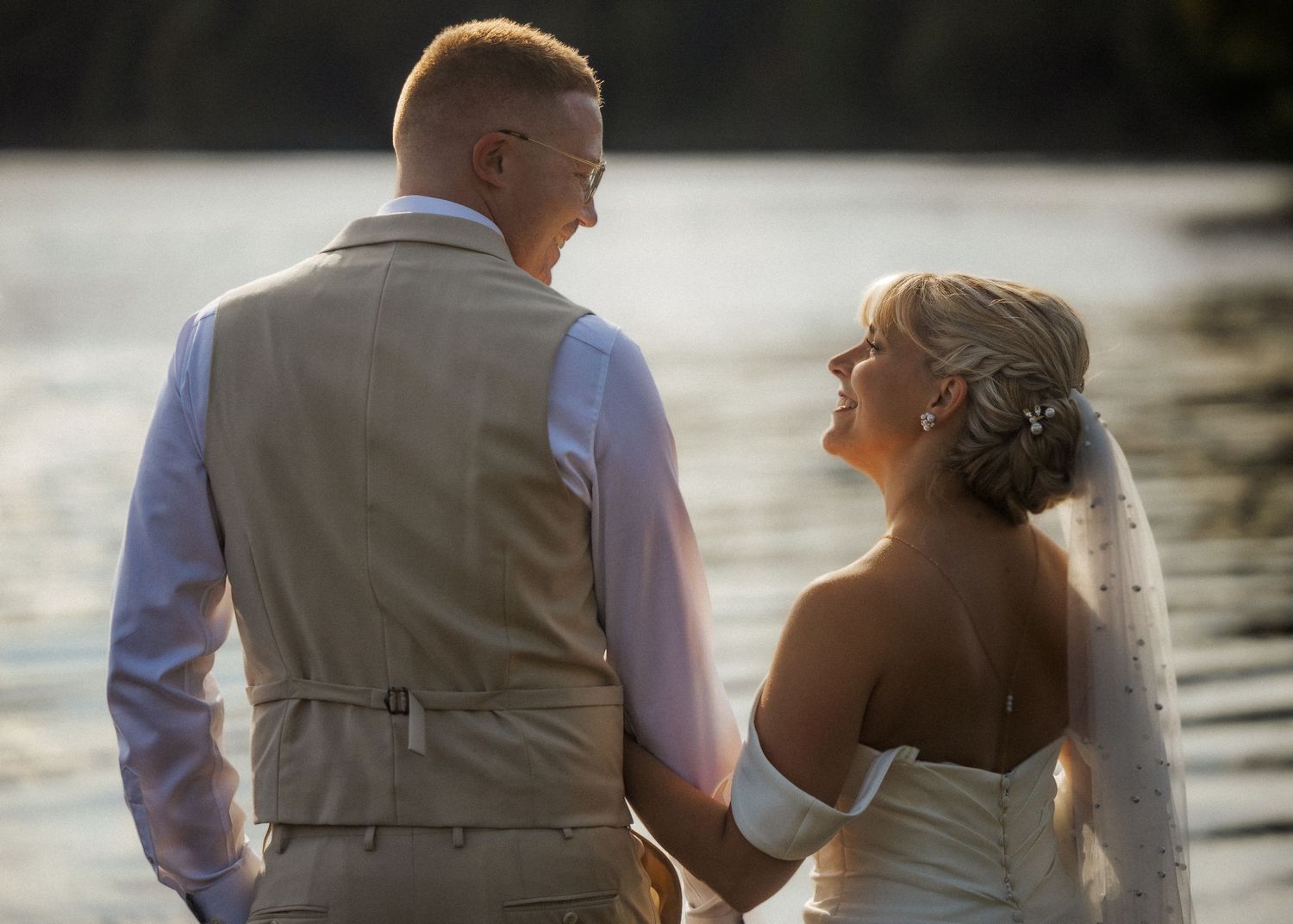 Couple in wedding attire standing together by the water at sunset.