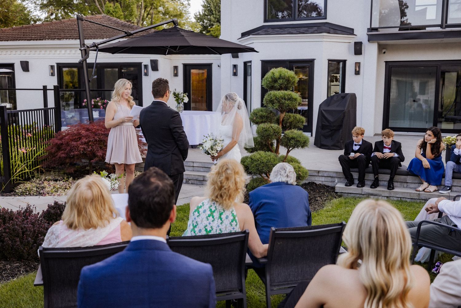 Outdoor wedding ceremony in front of modern white house with guests seated on lawn.