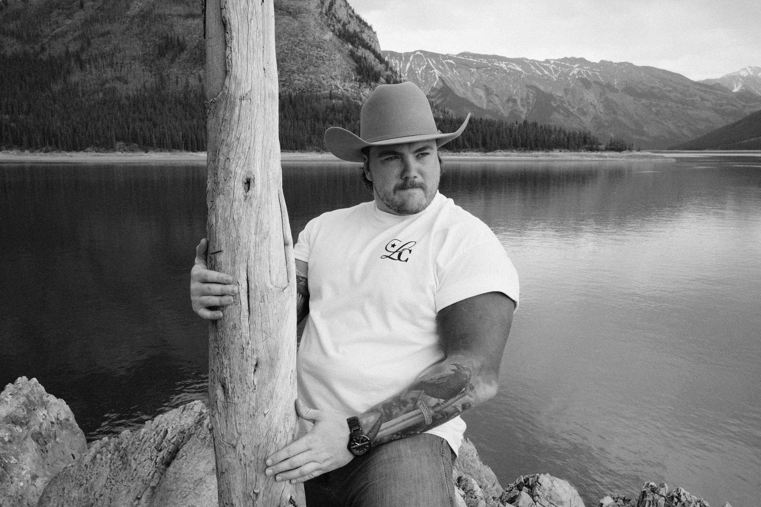 Man in cowboy hat leaning on wooden post by mountain lake, black and white outdoor portrait.