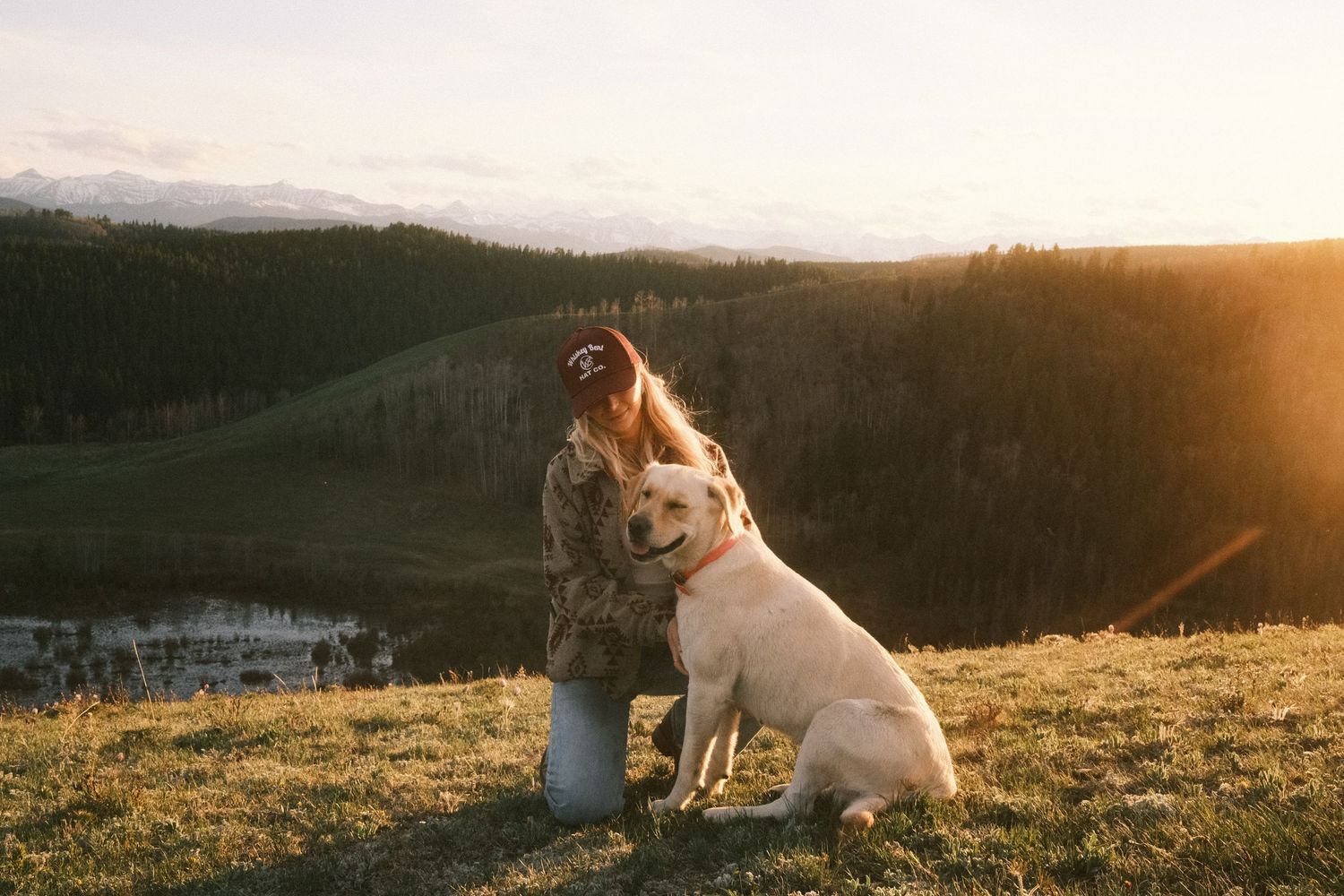 Woman kneeling with a yellow Labrador on a grassy hillside at golden sunset with mountain landscape.