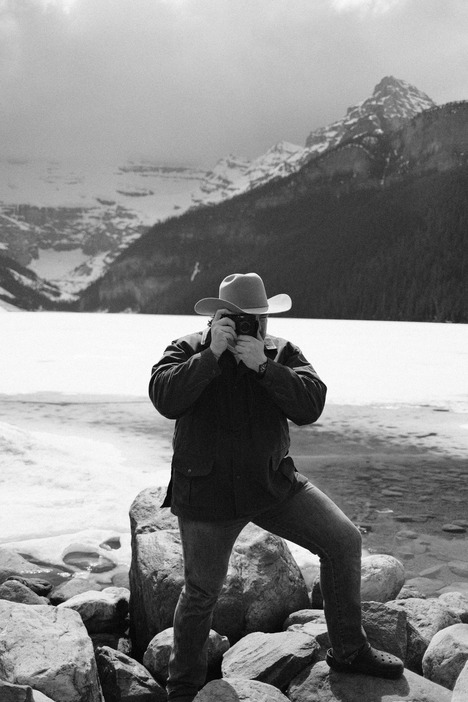 Black and white photo of a cowboy hat-wearing photographer shooting on snowy mountain rocks at Lake Louise.
