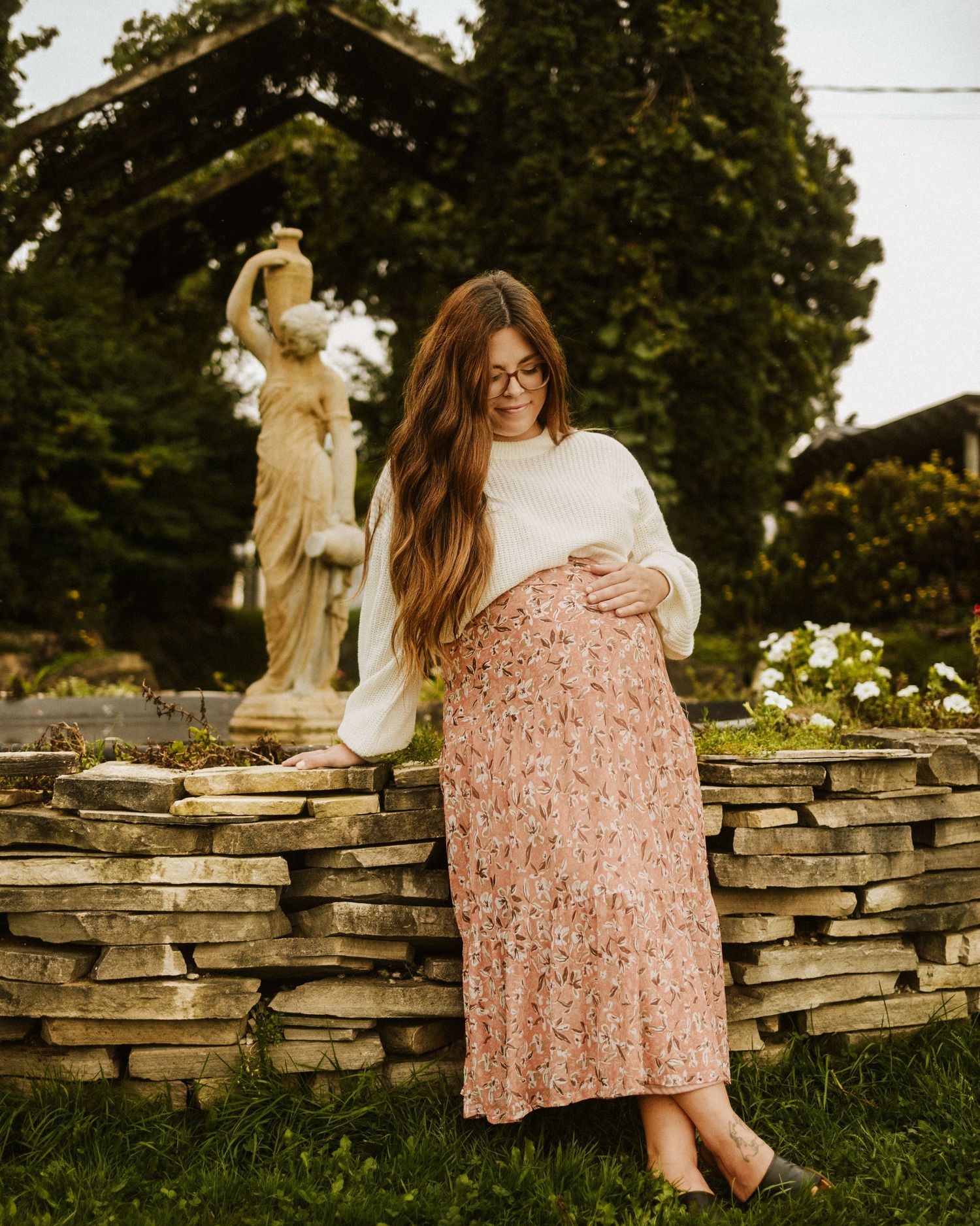 Person in floral dress sitting on rustic stone wall with garden statues.