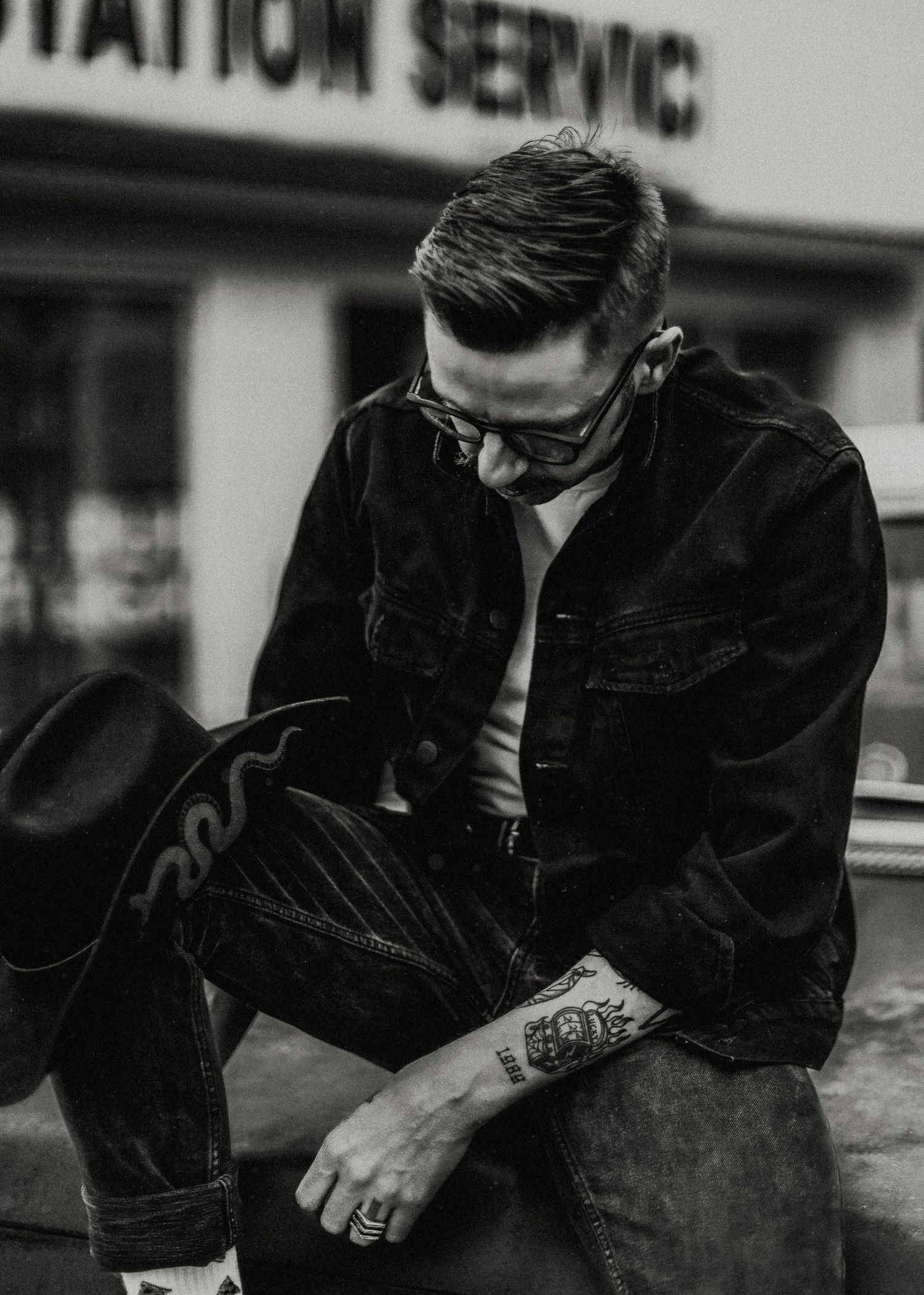 Person in denim jacket sitting at vintage service station in black and white.