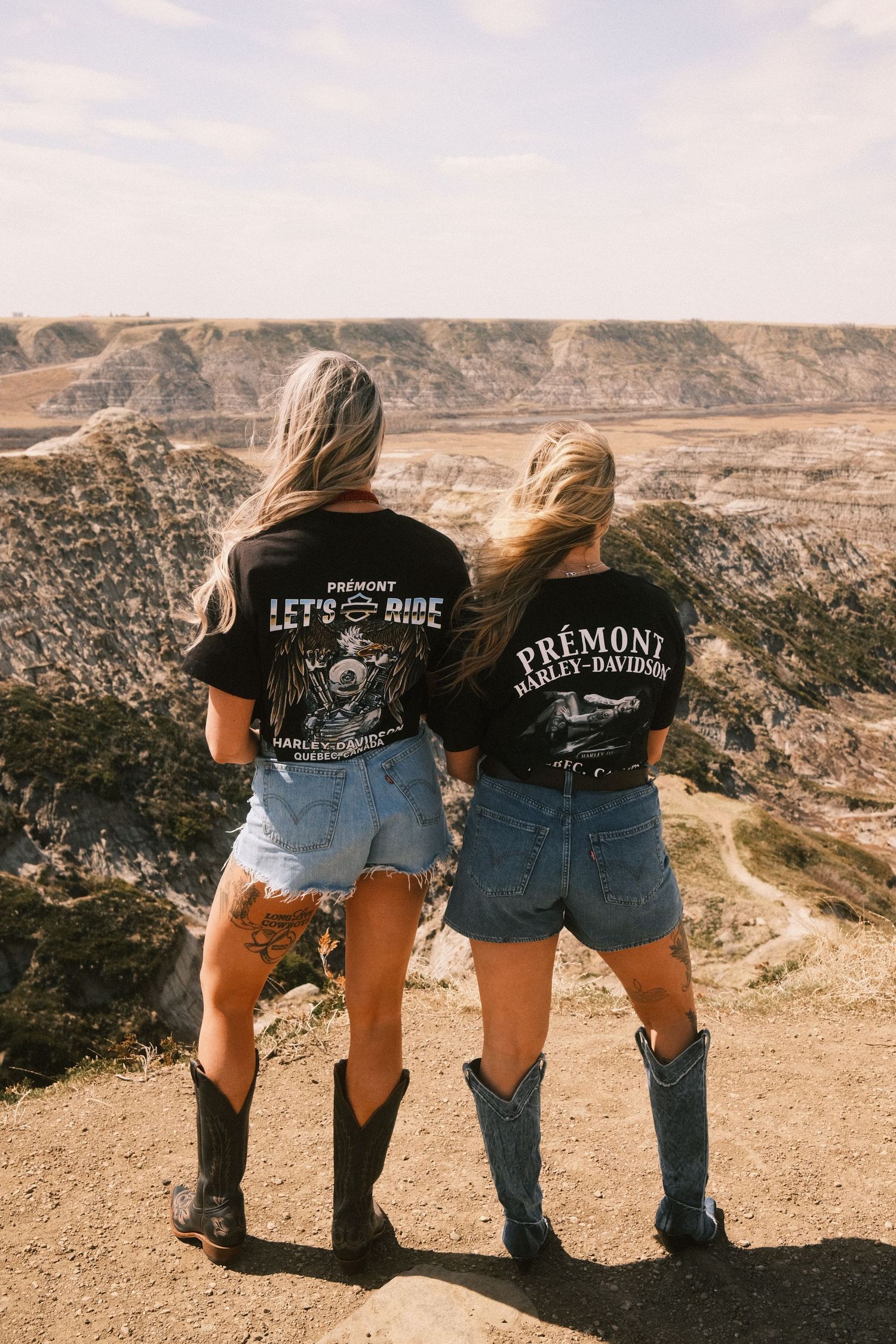 Two women in Harley-Davidson tees, denim shorts, and cowboy boots stand on a canyon overlook.