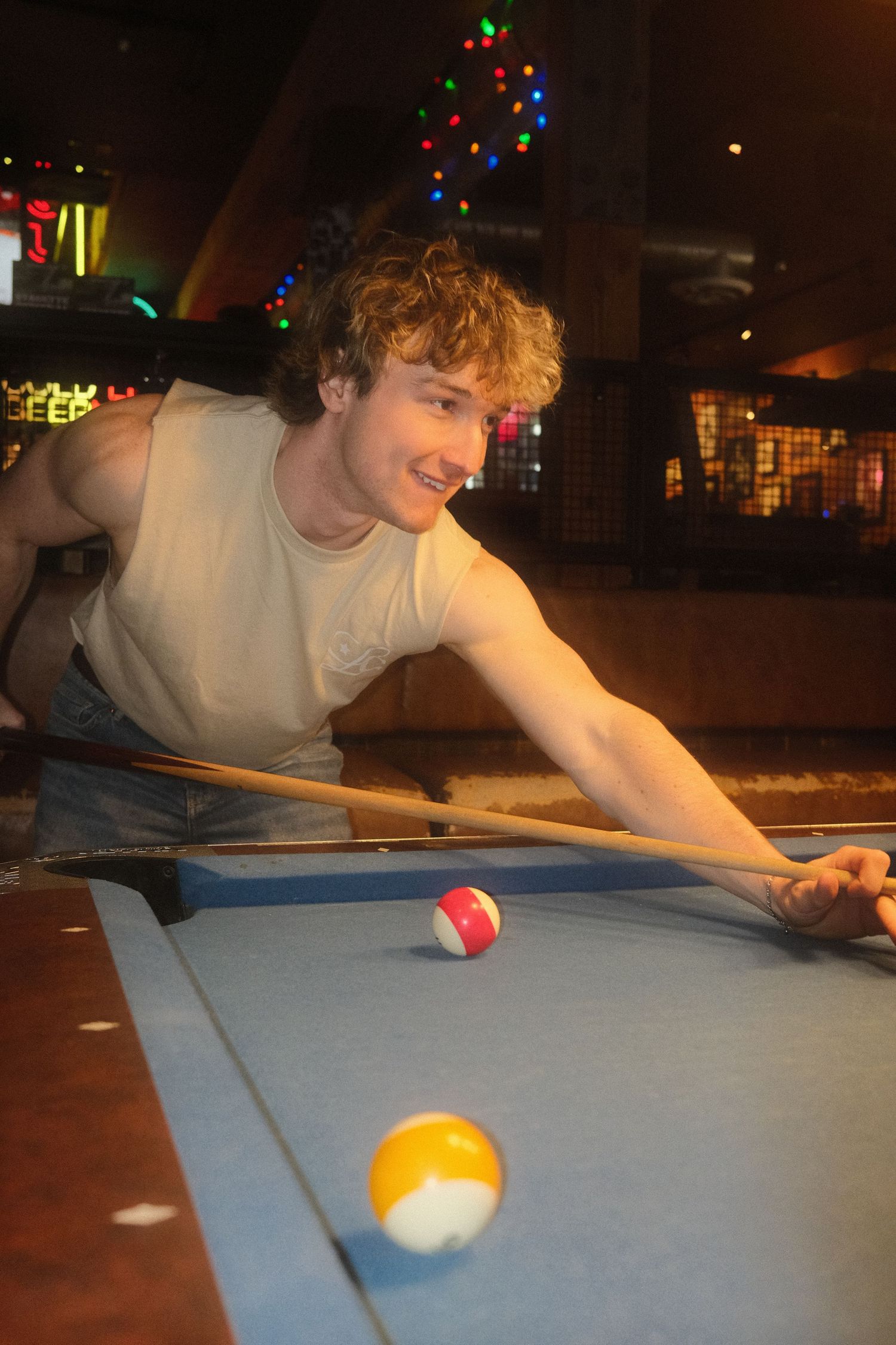 Young man with curly hair smiling while leaning over a pool table, holding a cue stick in a bar setting.