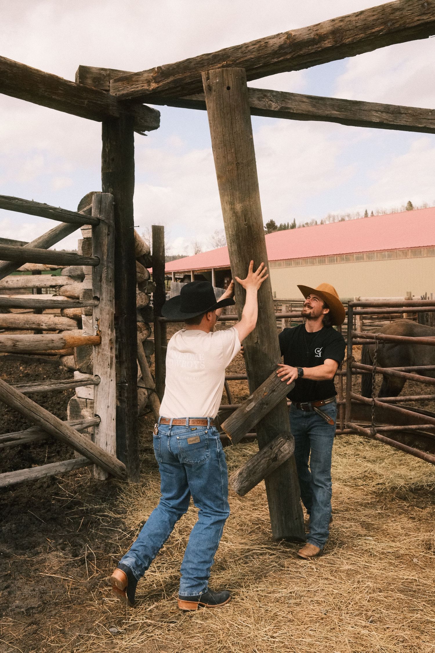 Two cowboys in hats and jeans work together on a rustic wooden fence at a ranch corral.