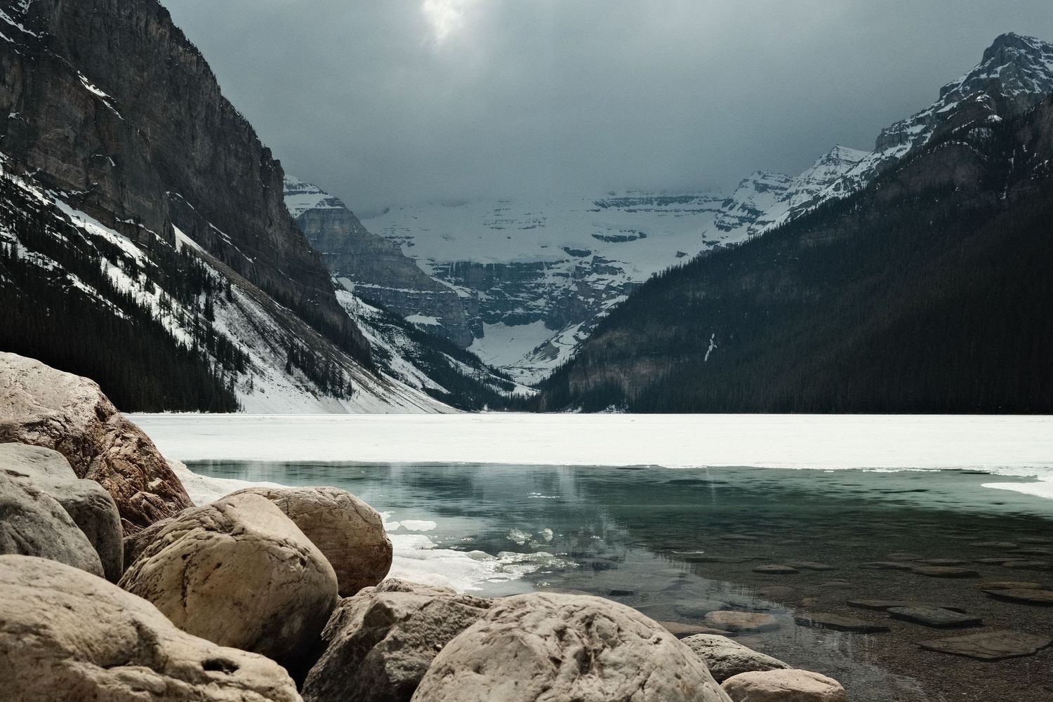 Snow-dusted rocky shoreline of Lake Louise with frozen turquoise water and dramatic mountain peaks under stormy winter skies.
