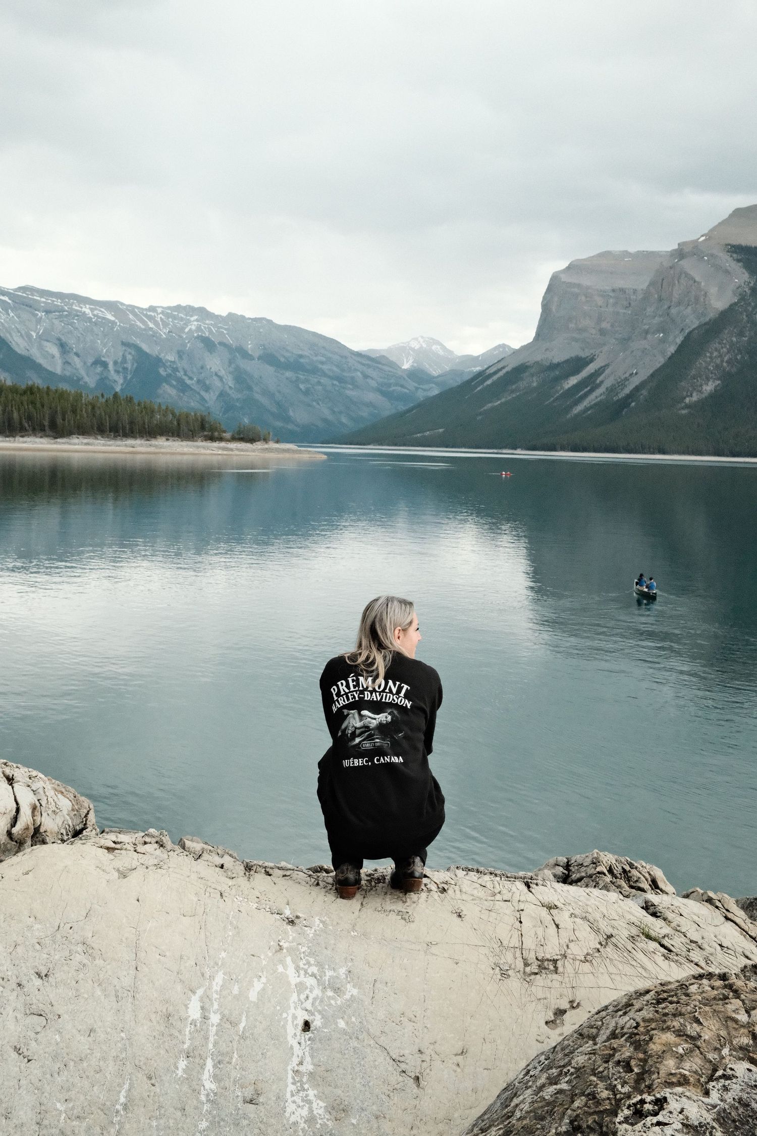Woman in black Fremont hoodie crouching on rocky shore of serene mountain lake with snow-capped peaks.