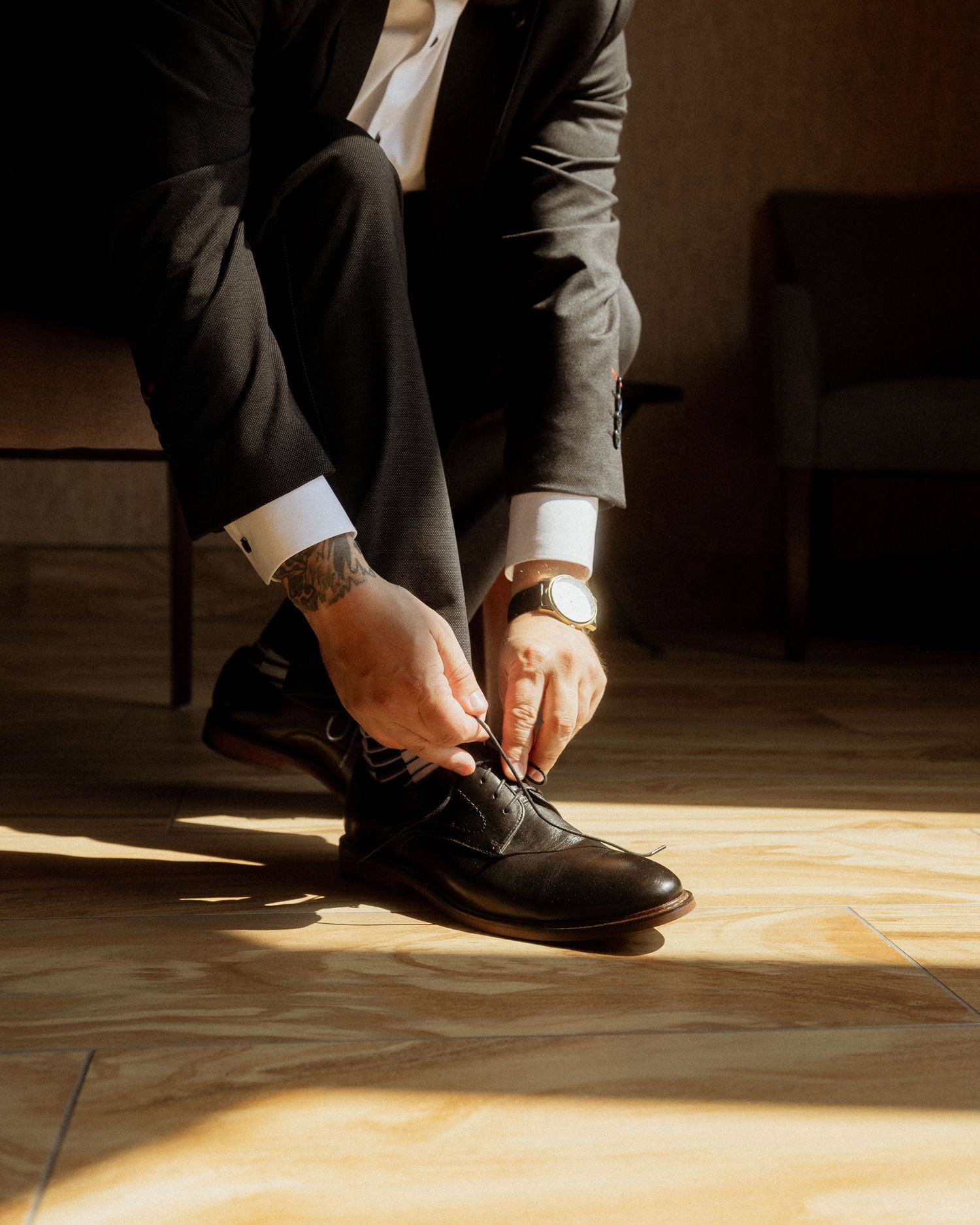 Close-up shot of hands adjusting formal black dress shoes on wooden floor in dramatic lighting.