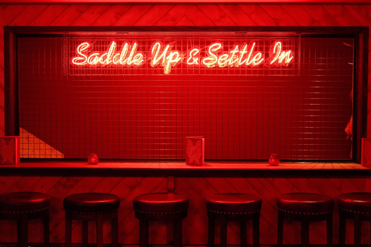 Red neon sign reading Saddle Up & Settle In above a bar counter with dark wooden stools in moody red lighting.