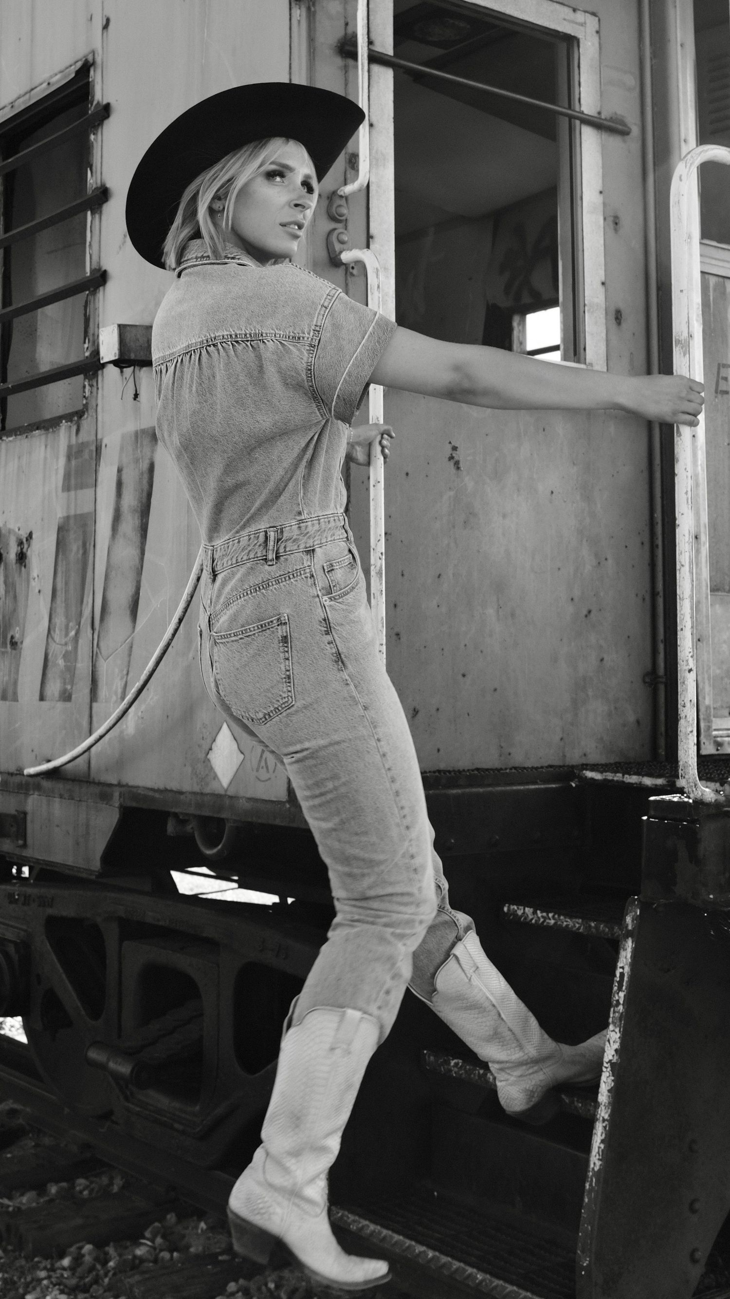 Black and white photo of a blonde woman in cowboy hat, denim outfit, and white boots posing on vintage train car steps.